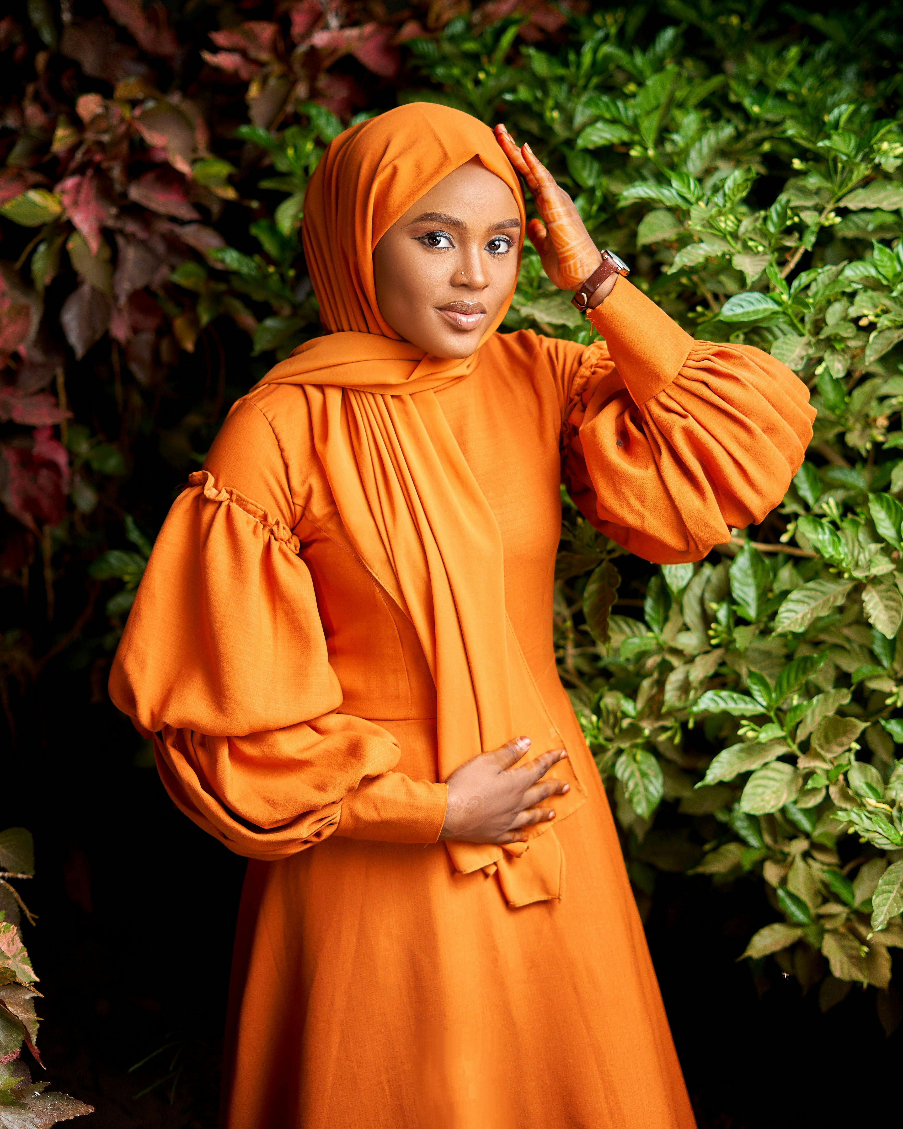 Portrait of an elegant woman in an orange abaya surrounded by lush greenery.