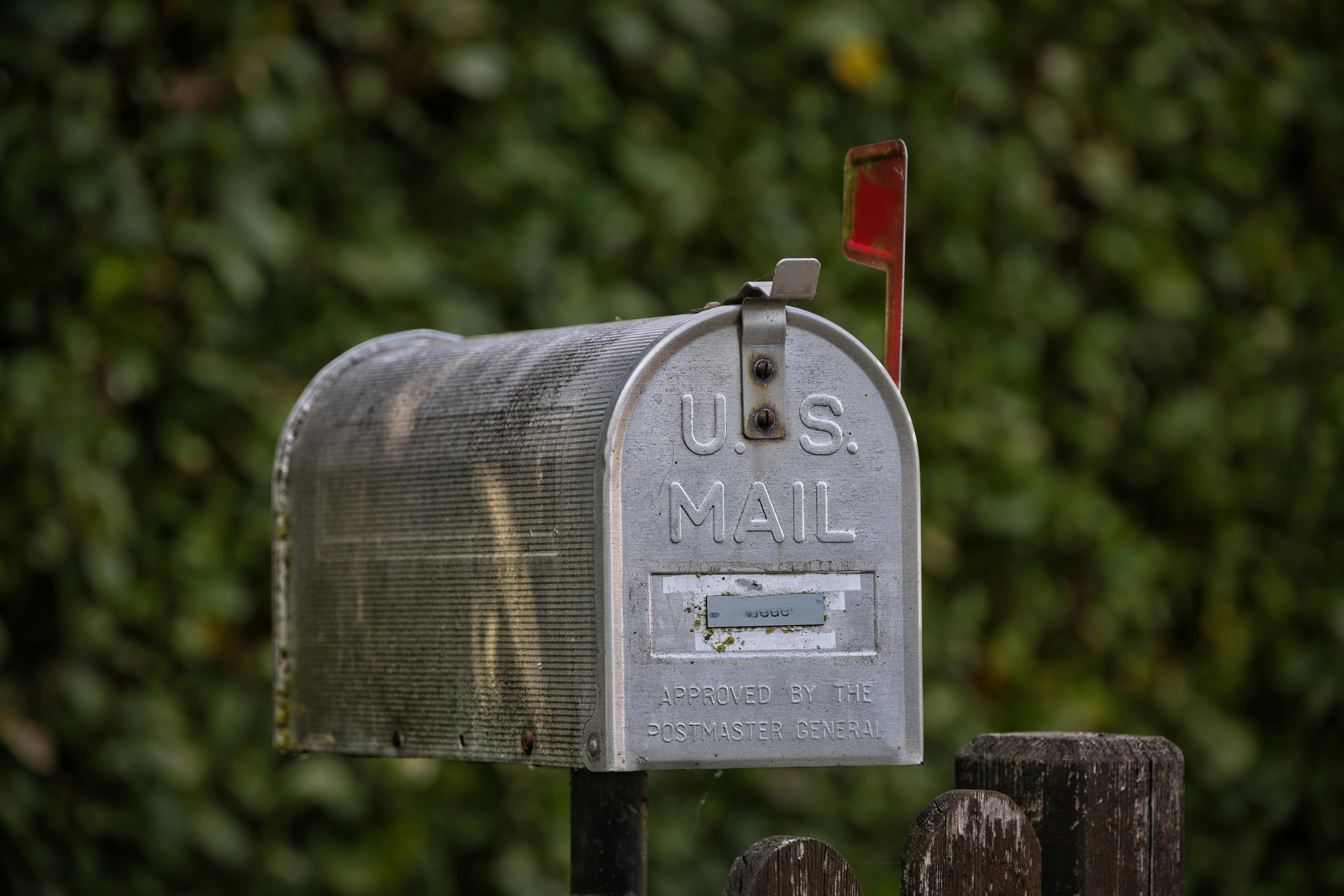 A traditional U.S. mailbox with red flag against a lush green blurred background. | BocaVibe