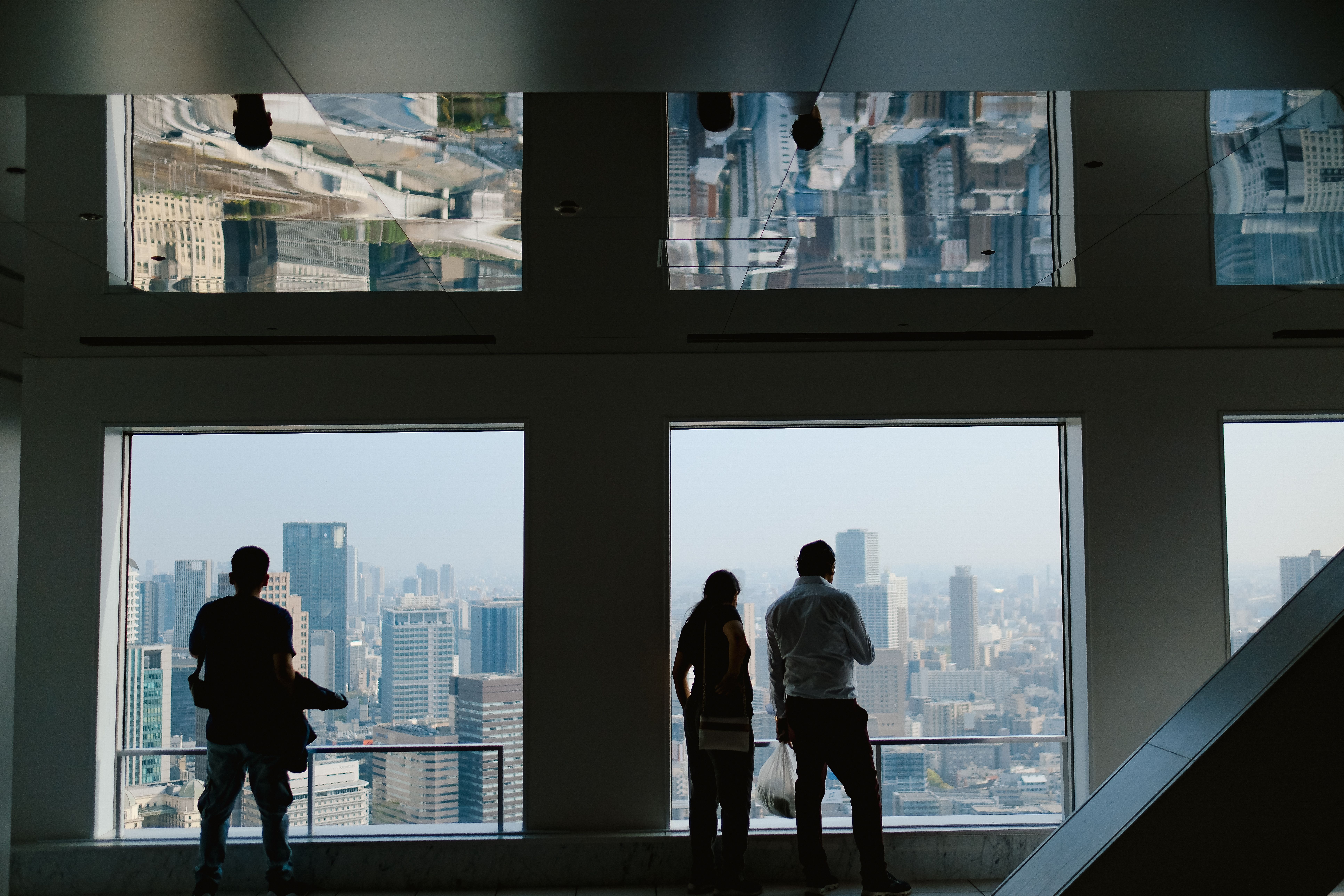 Silhouettes admire a city skyline through windows indoors, reflecting urban architecture.