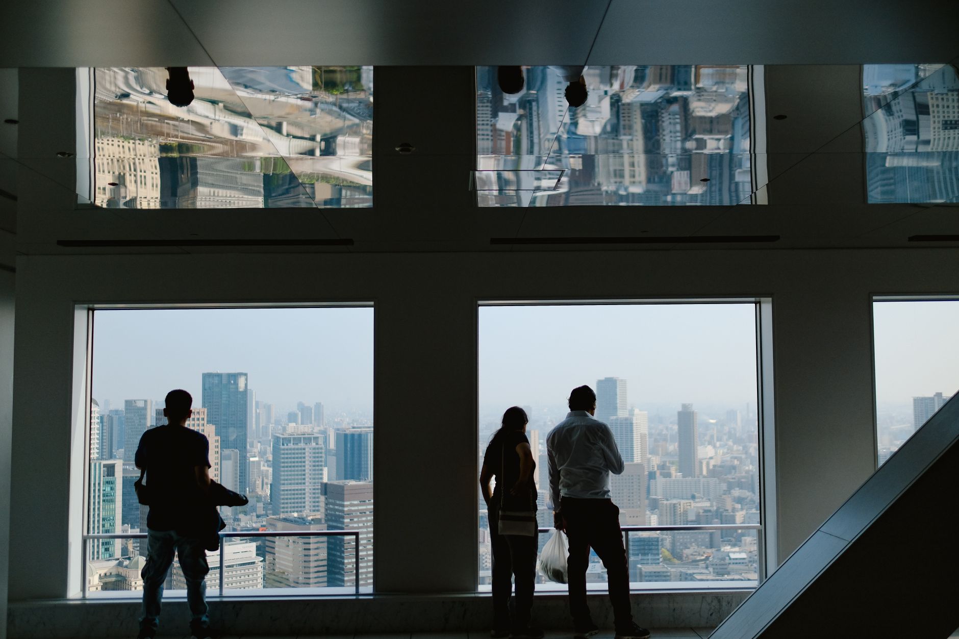 Silhouettes admire a city skyline through windows indoors, reflecting urban architecture.