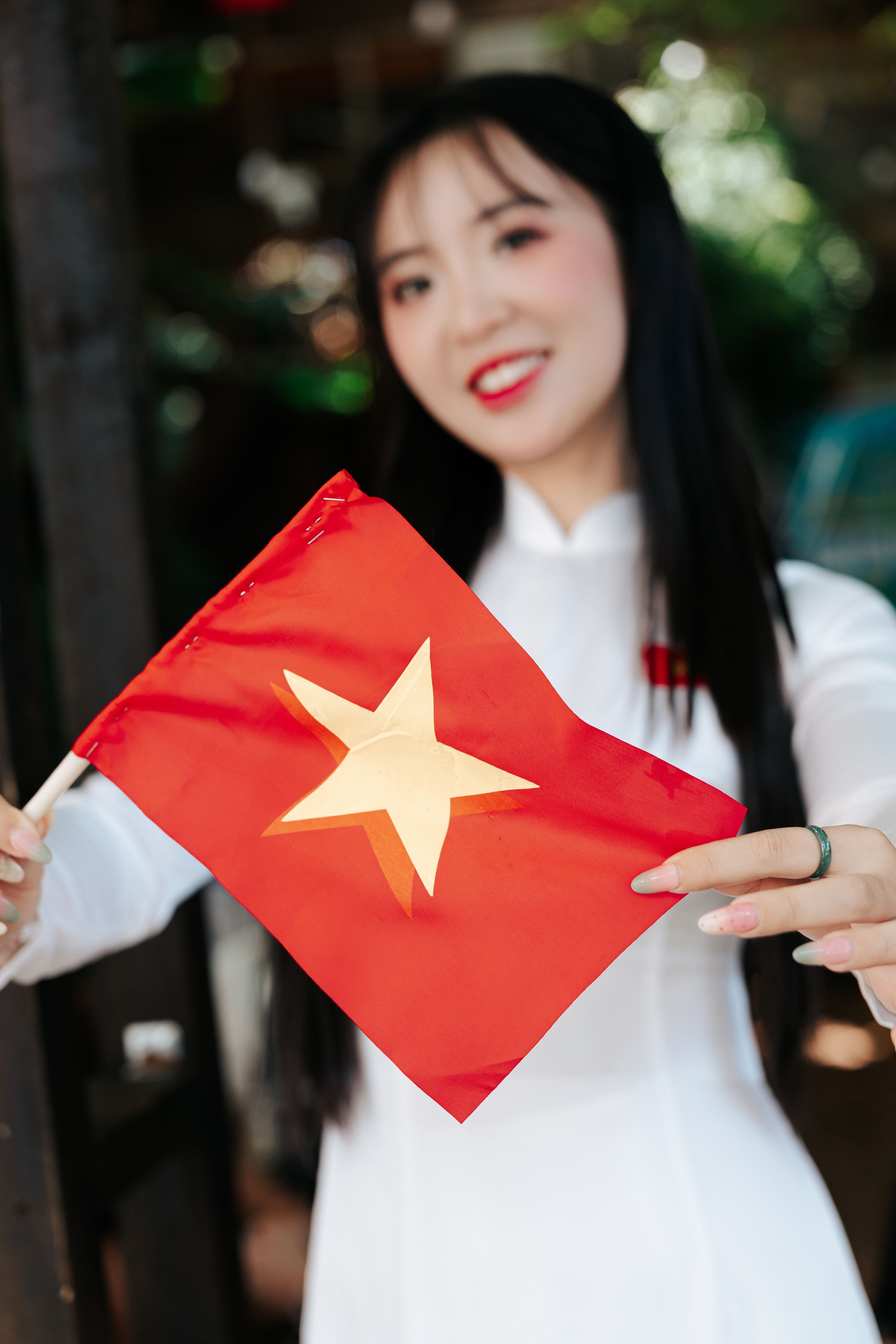 Smiling woman in traditional dress holding a Vietnamese flag, outdoors.