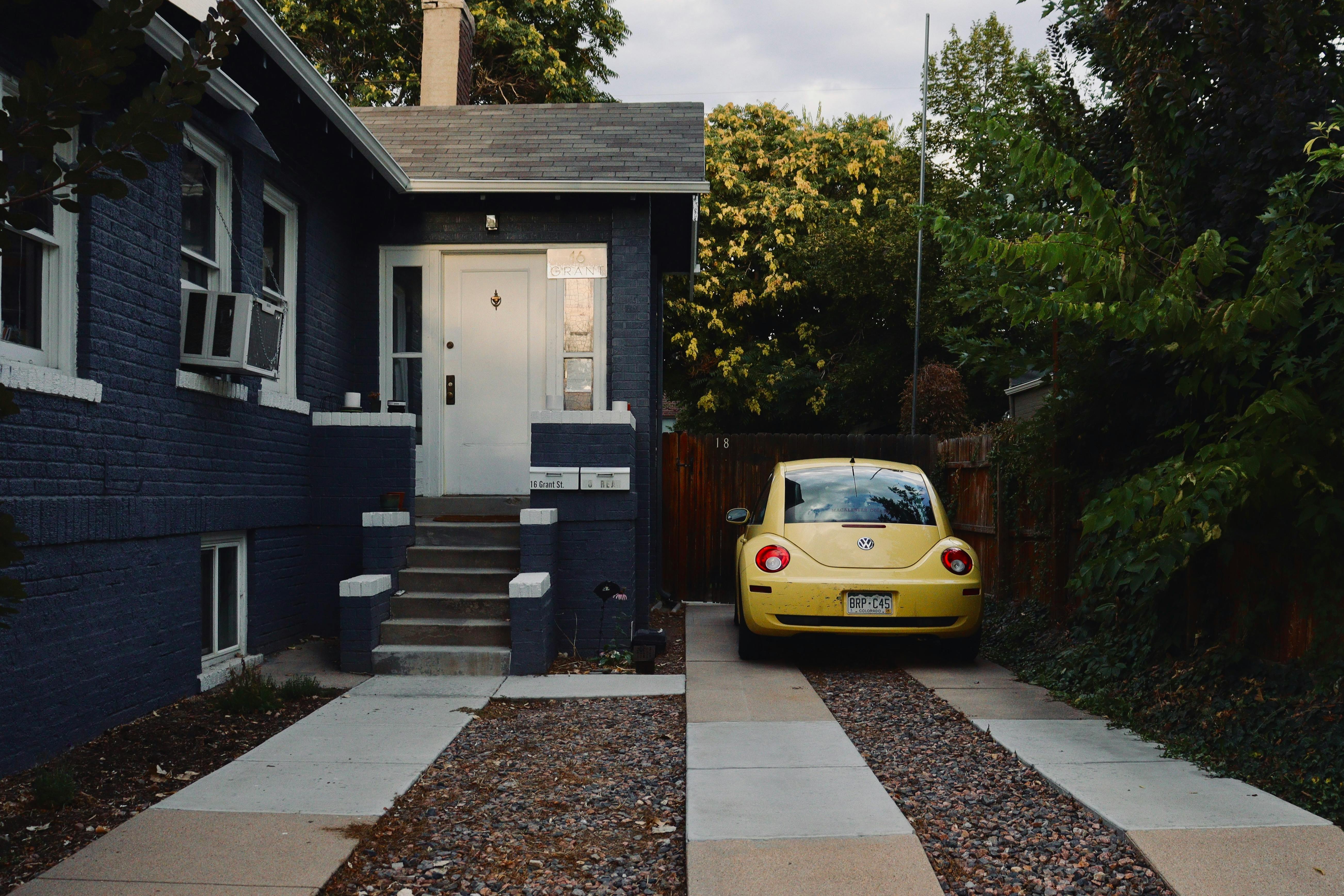 Yellow Volkswagen Beetle parked in a driveway beside a dark blue brick house.