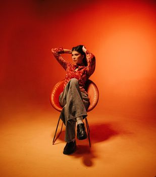 Stylish woman posing in a modern red chair with an orange background, exuding confidence and elegance.
