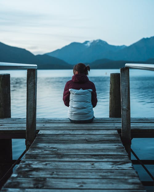 Woman Wearing White and Marron Jacket Sitting on Brown Wooedn Boardwalk