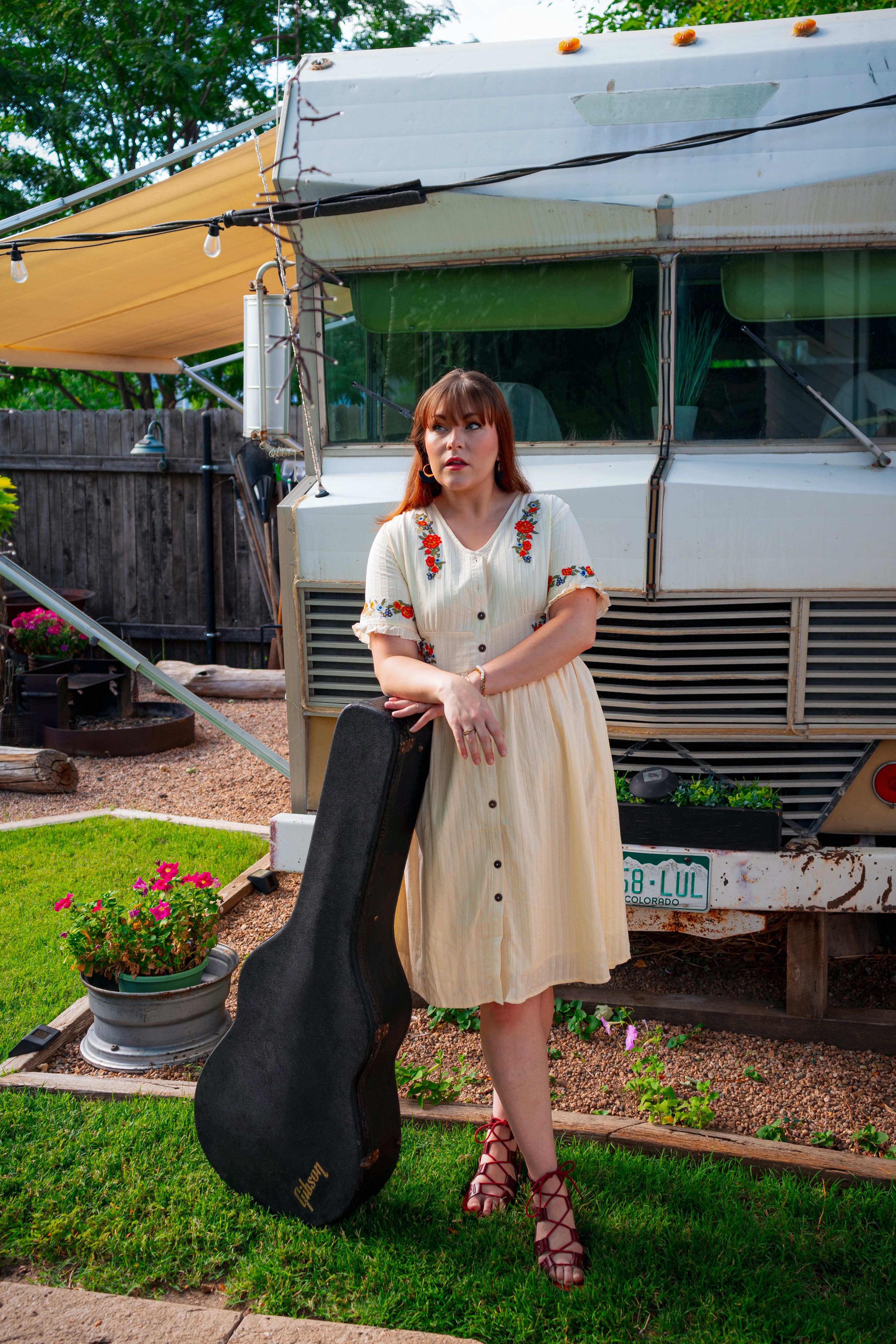 Woman in vintage dress with guitar case outdoors by a retro RV in summer.