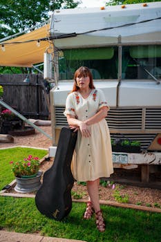 Woman in vintage dress with guitar case outdoors by a retro RV in summer.