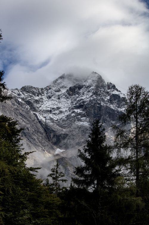 Free Majestic snow-covered mountain peak surrounded by lush forest under a cloudy sky. Stock Photo
