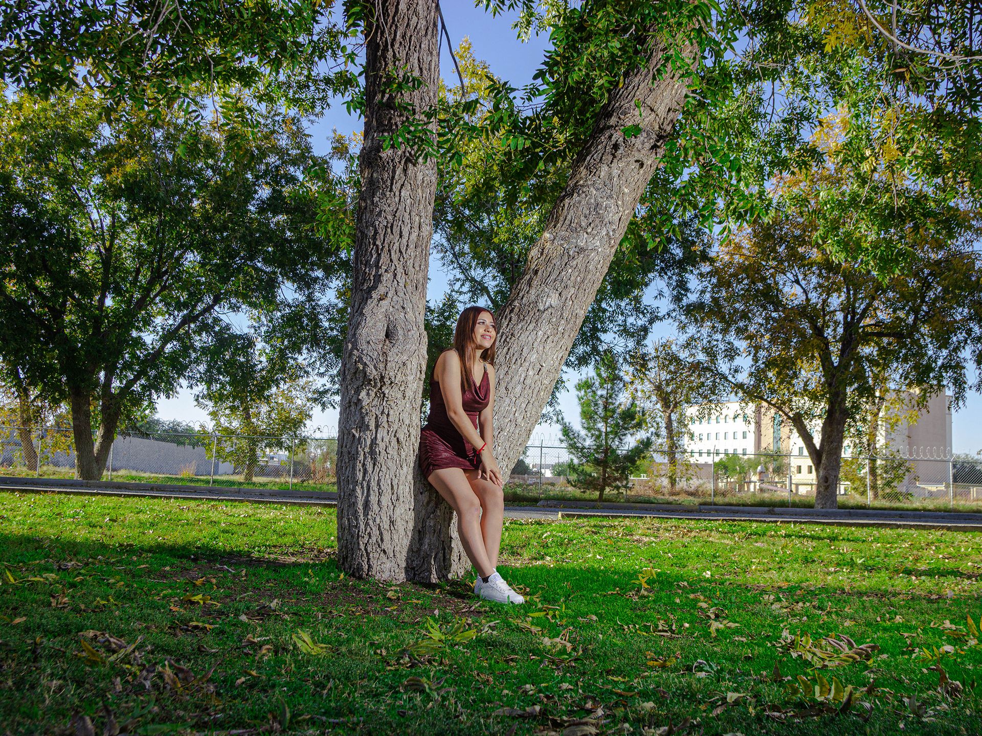 Young woman in a burgundy dress sits under a tree in a sunny, urban park setting.