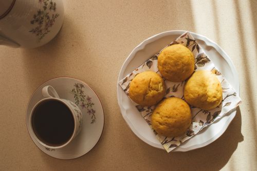 Free Warm morning light bathes a cup of coffee and muffins on a floral plate. Stock Photo