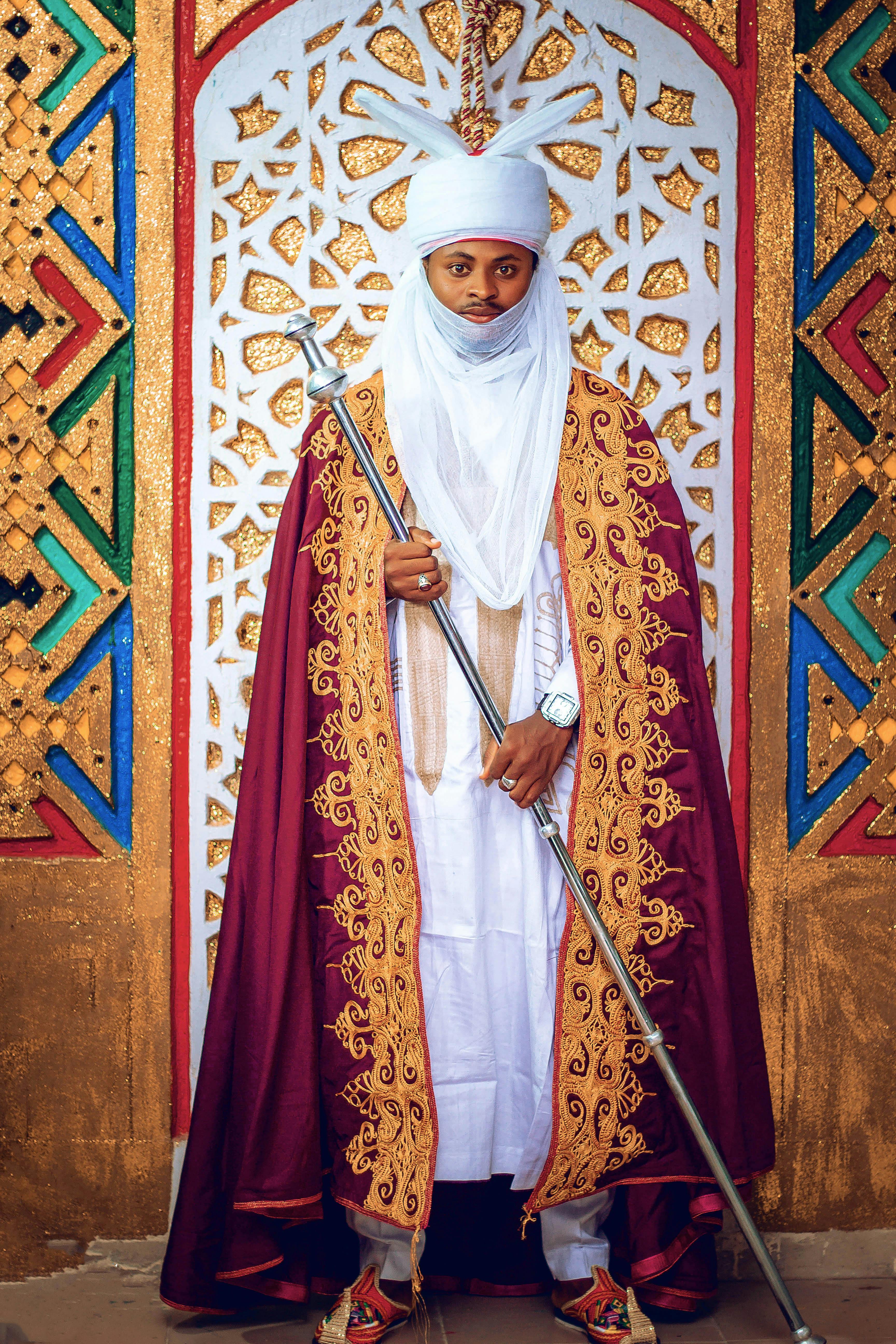 A man in traditional African attire stands in front of a colorful ornate backdrop holding a staff.