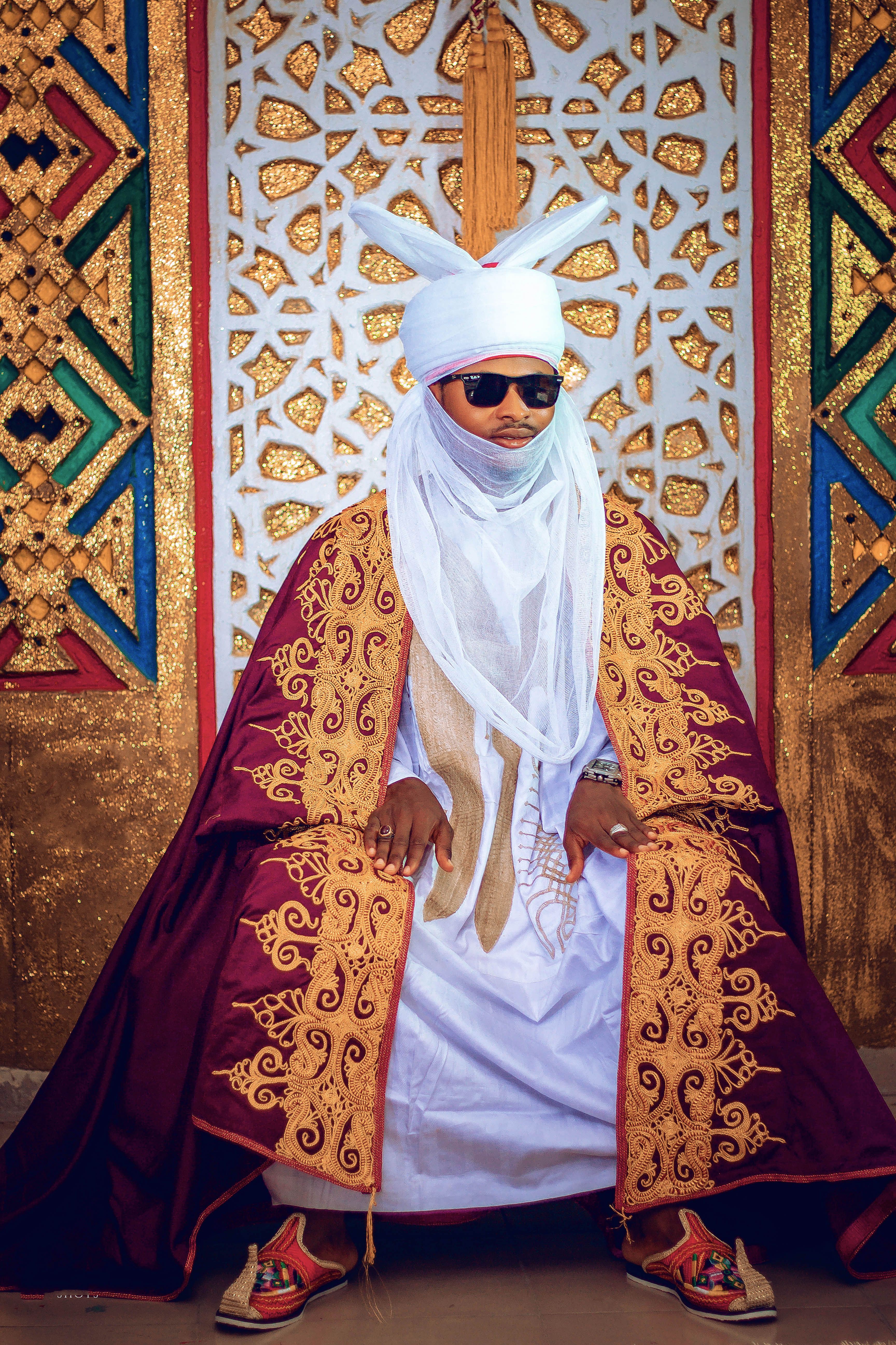 Man in traditional attire seated against intricate patterned backdrop, exuding cultural elegance.
