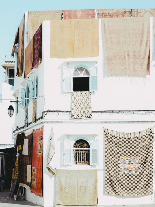 Free White building in Asilah, Morocco, adorned with traditional patterned rugs hanging outdoors. Stock Photo