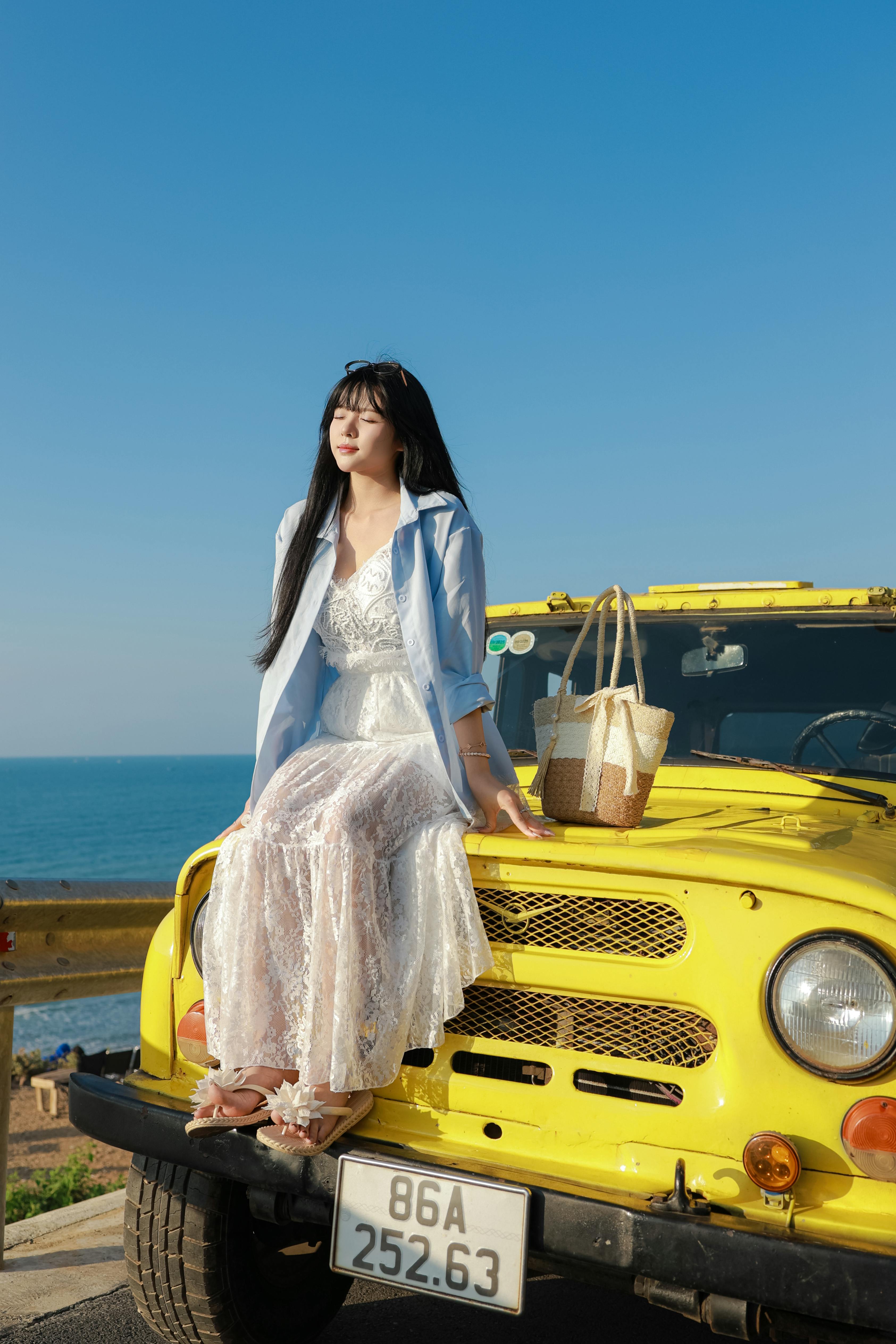 A stylish woman in a white dress sits on a yellow jeep, enjoying a sunny coastal day.