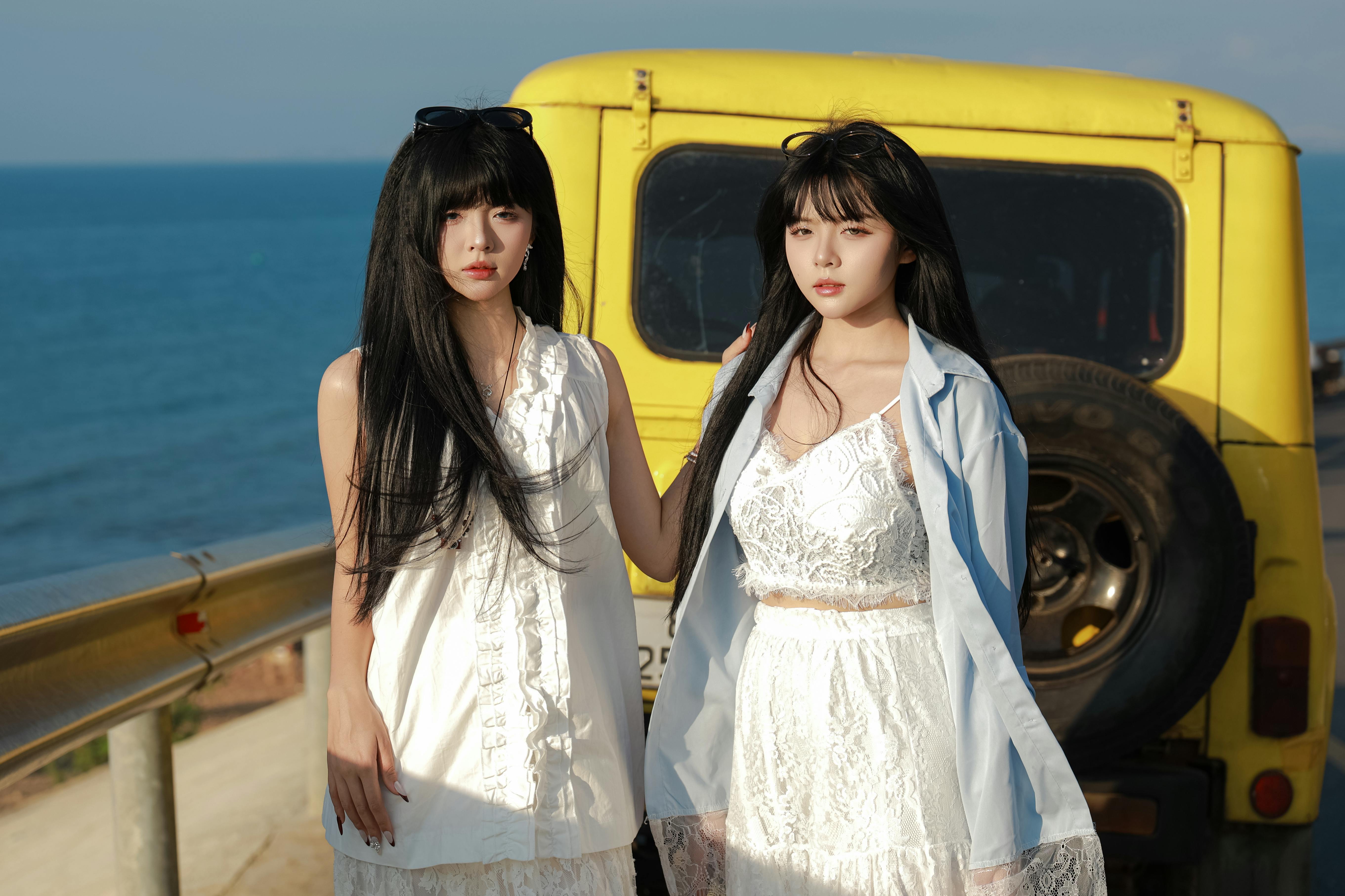 Two women in white dresses pose by a yellow car near the ocean on a sunny day.