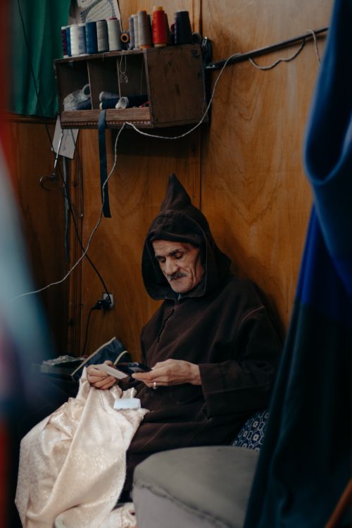 Free Craftsman in hooded robe focuses on sewing indoors, surrounded by threads. Stock Photo