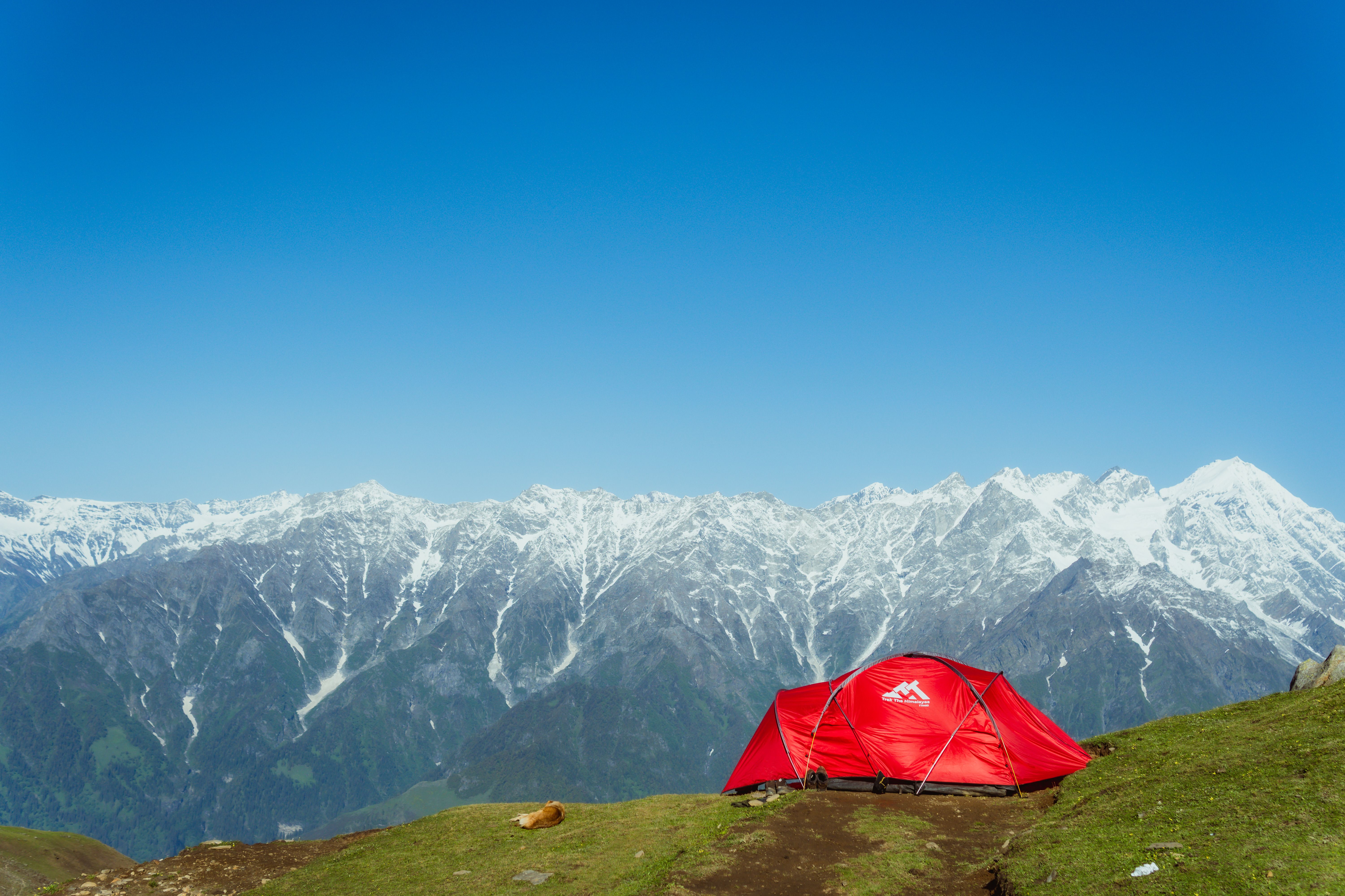 Red camping tent on a grassy hilltop with majestic snowy mountains in the background and clear blue sky.