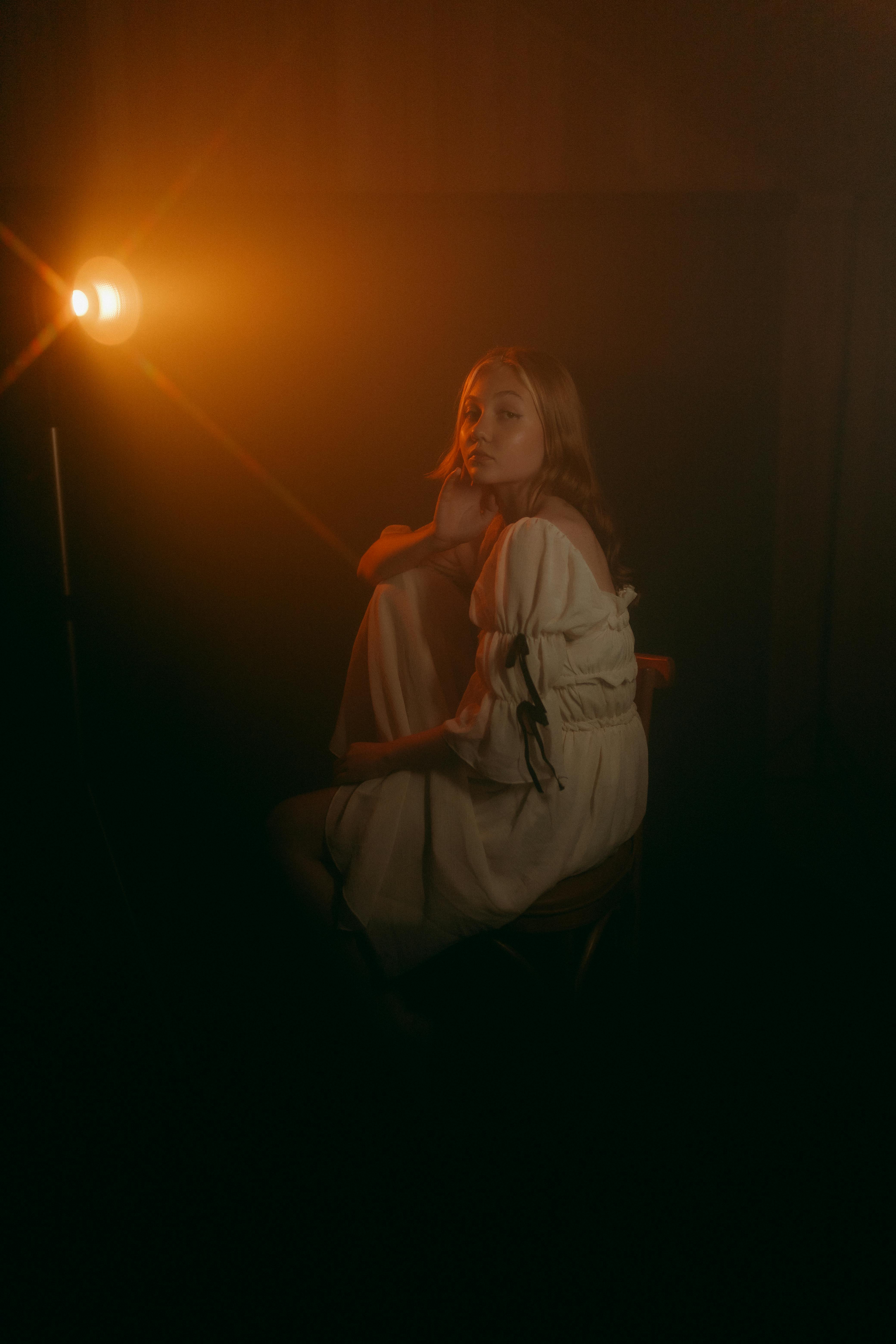 Ücretsiz Sıcak ve dramatik bir ışıklandırma altında düşünceli bir şekilde oturan bir kadın, kasvetli bir portre atmosferi yaratıyor. Stok Fotoğraflar