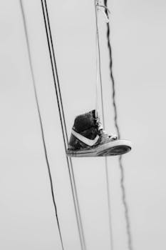 Monochrome image of a single sneaker hanging by laces on cables, symbolizing urban street culture.
