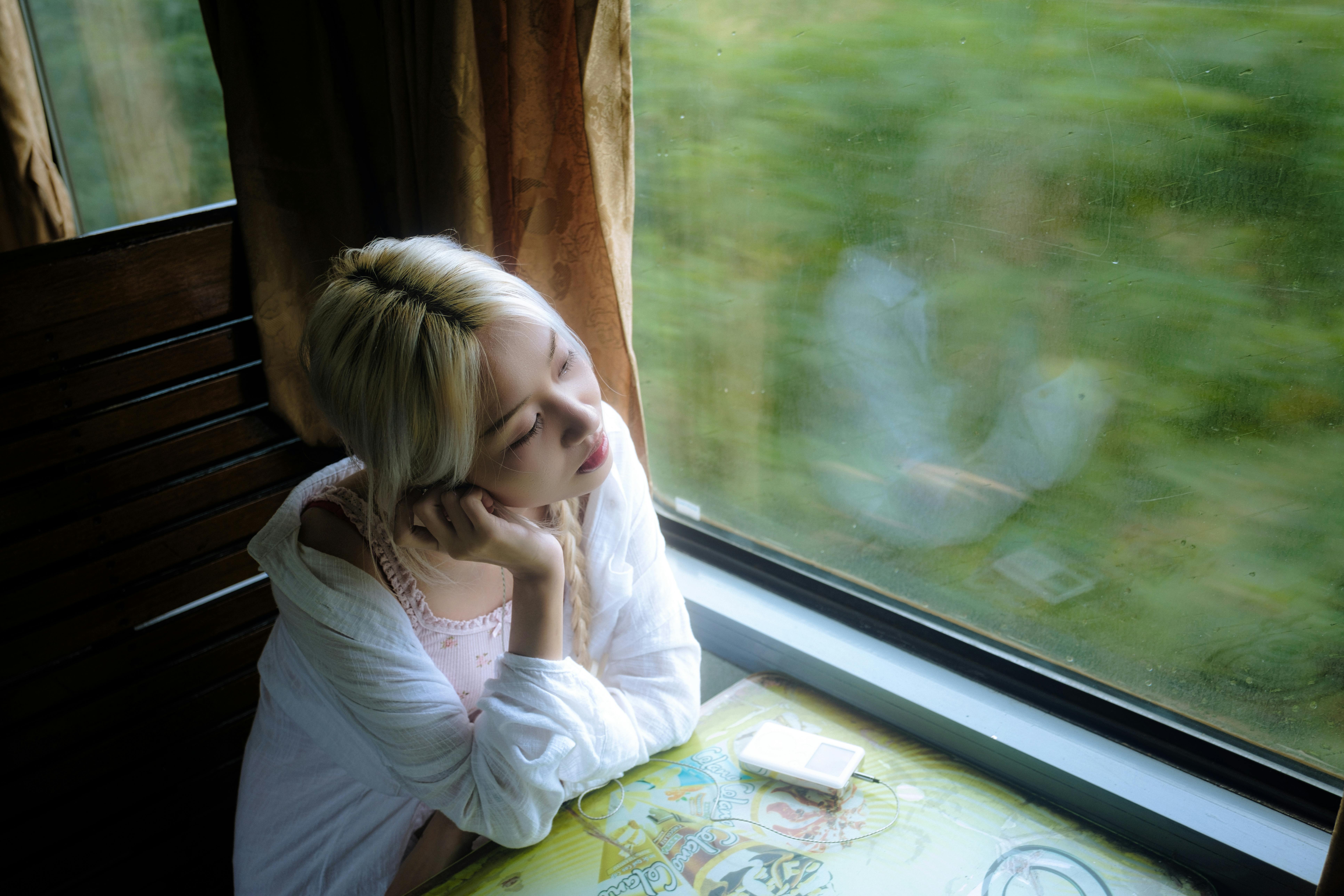 A woman with blonde hair in a white outfit gazes out a train window, reflecting nature.