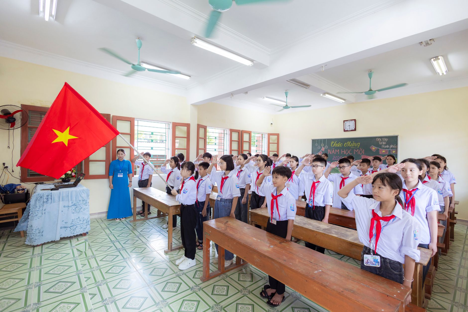 Students in uniform saluting the Vietnamese flag during a school ceremony in a classroom.