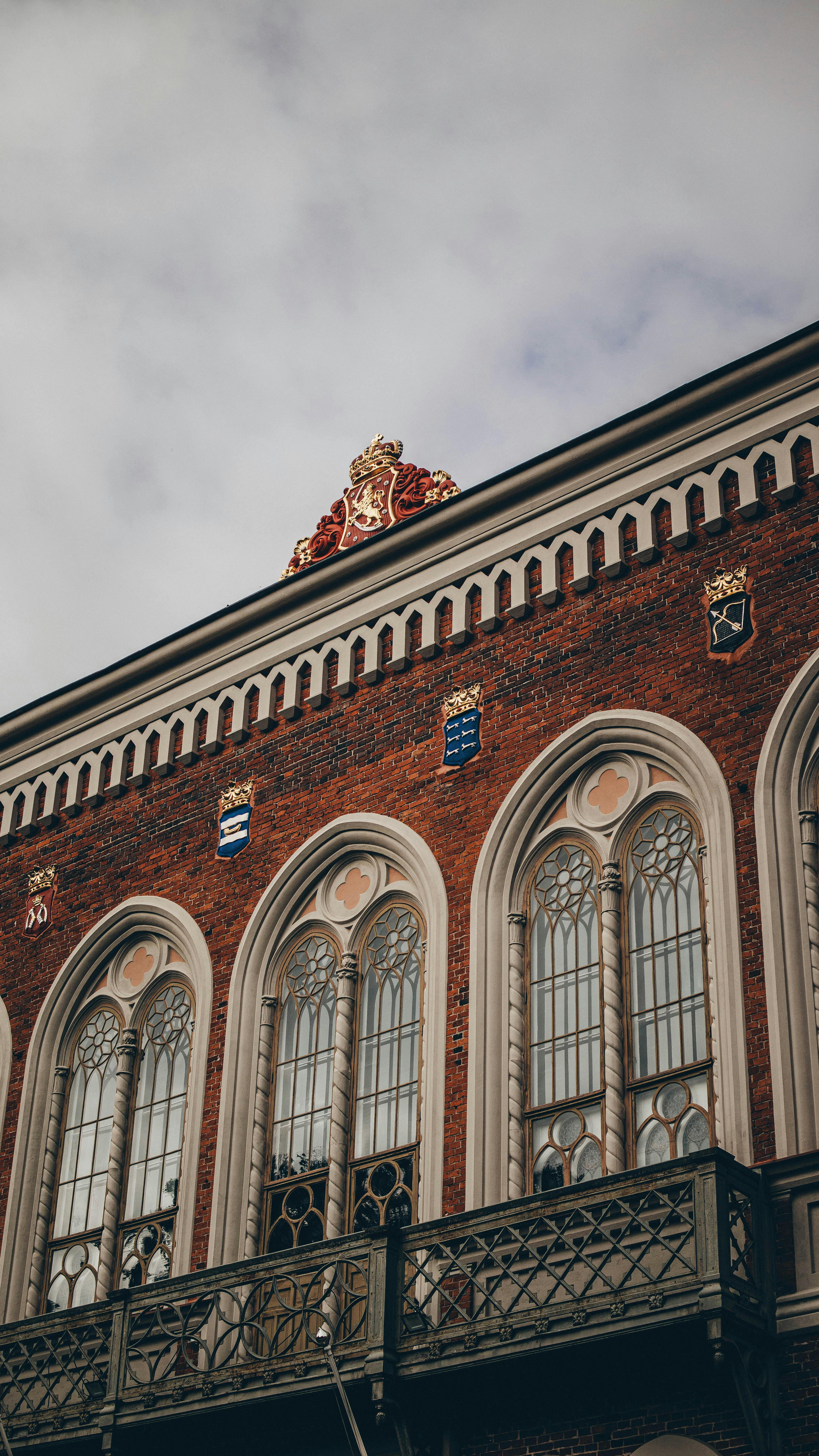 Gratis Elegante fachada histórica de edificio de ladrillo con ventanas decorativas y escudos de armas bajo un cielo nublado. Foto de stock