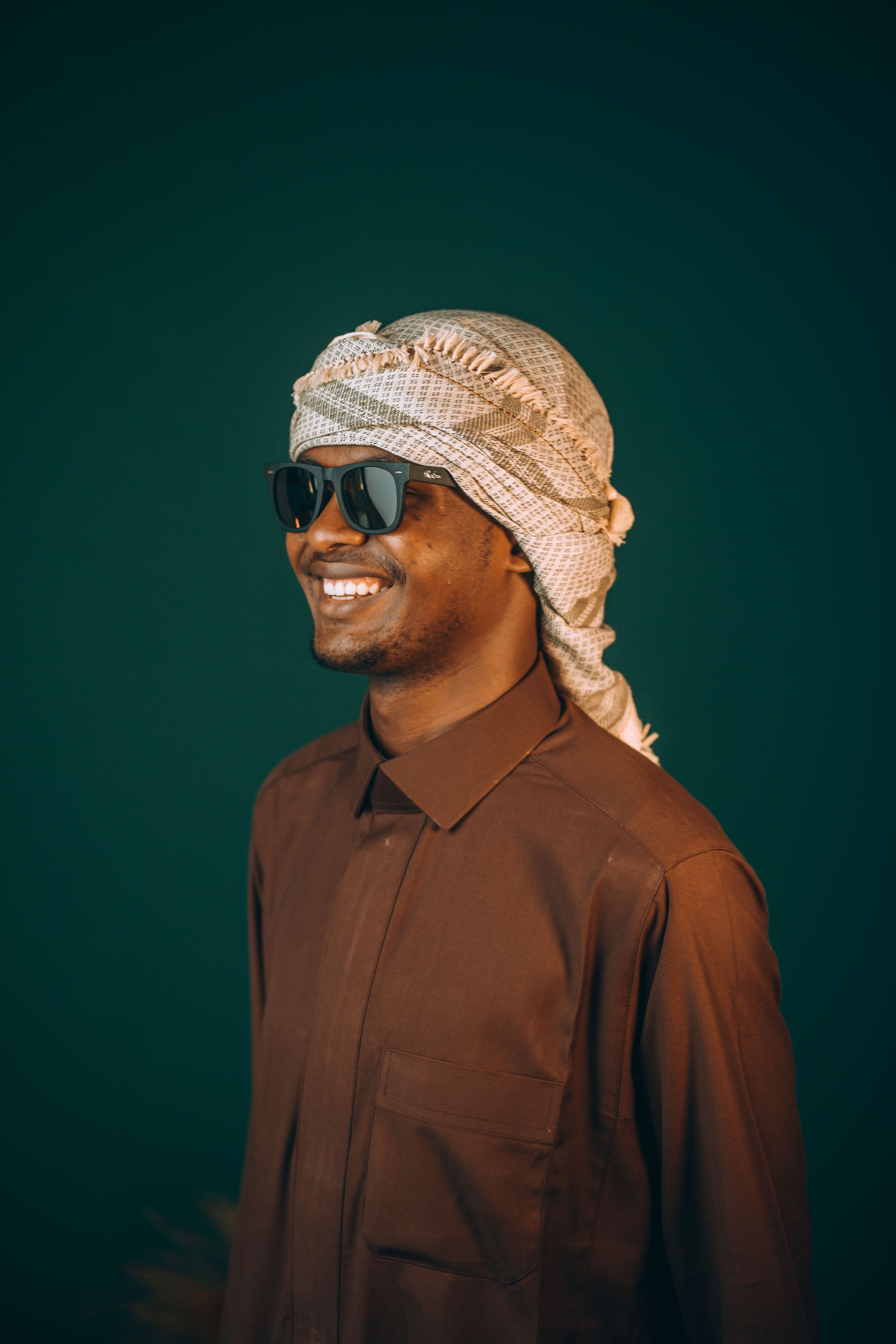 Smiling man in traditional Middle Eastern attire and sunglasses against a green background, Dubai setting.