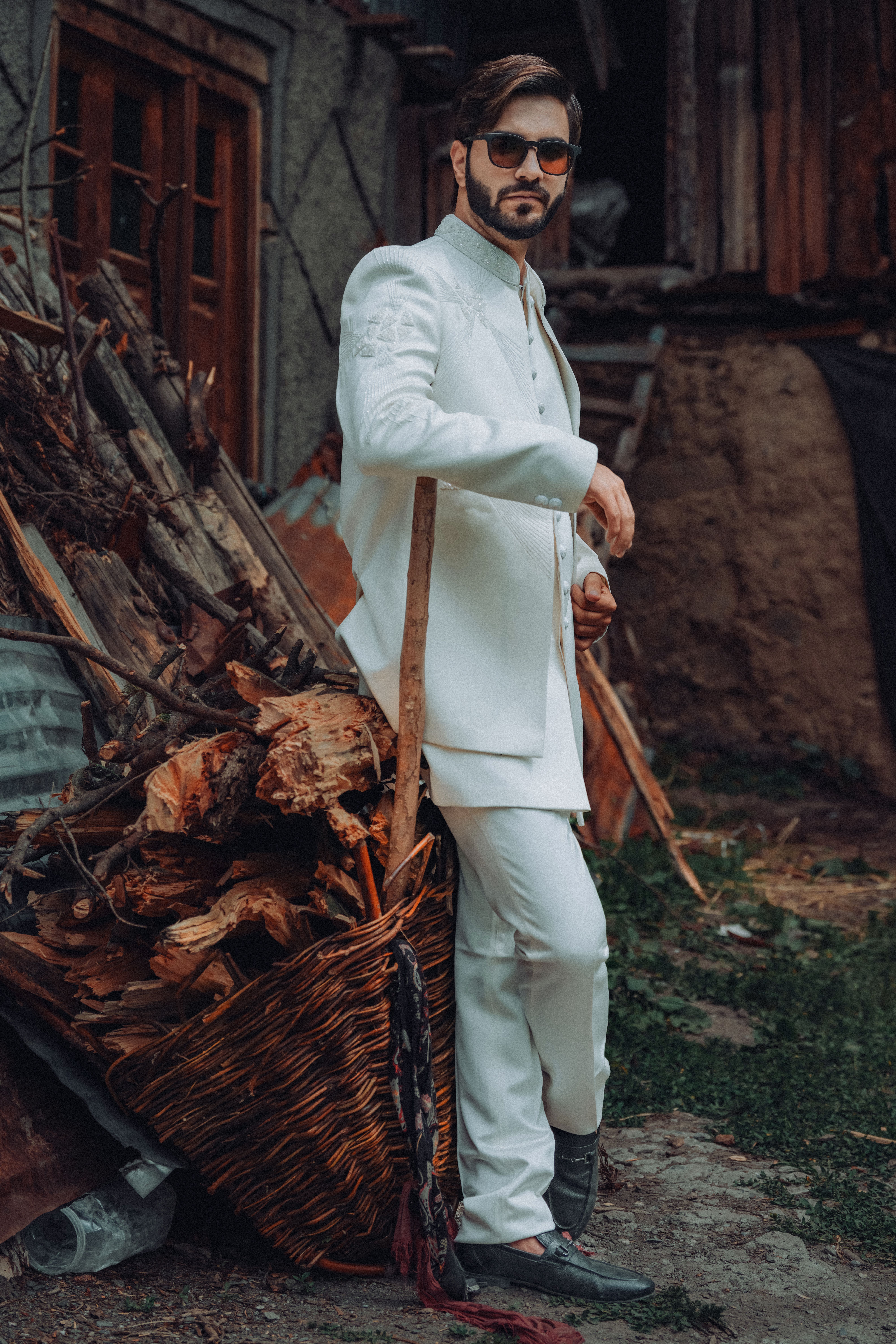 A fashionable man poses confidently in a white suit against a rustic backdrop, exuding style and charm.