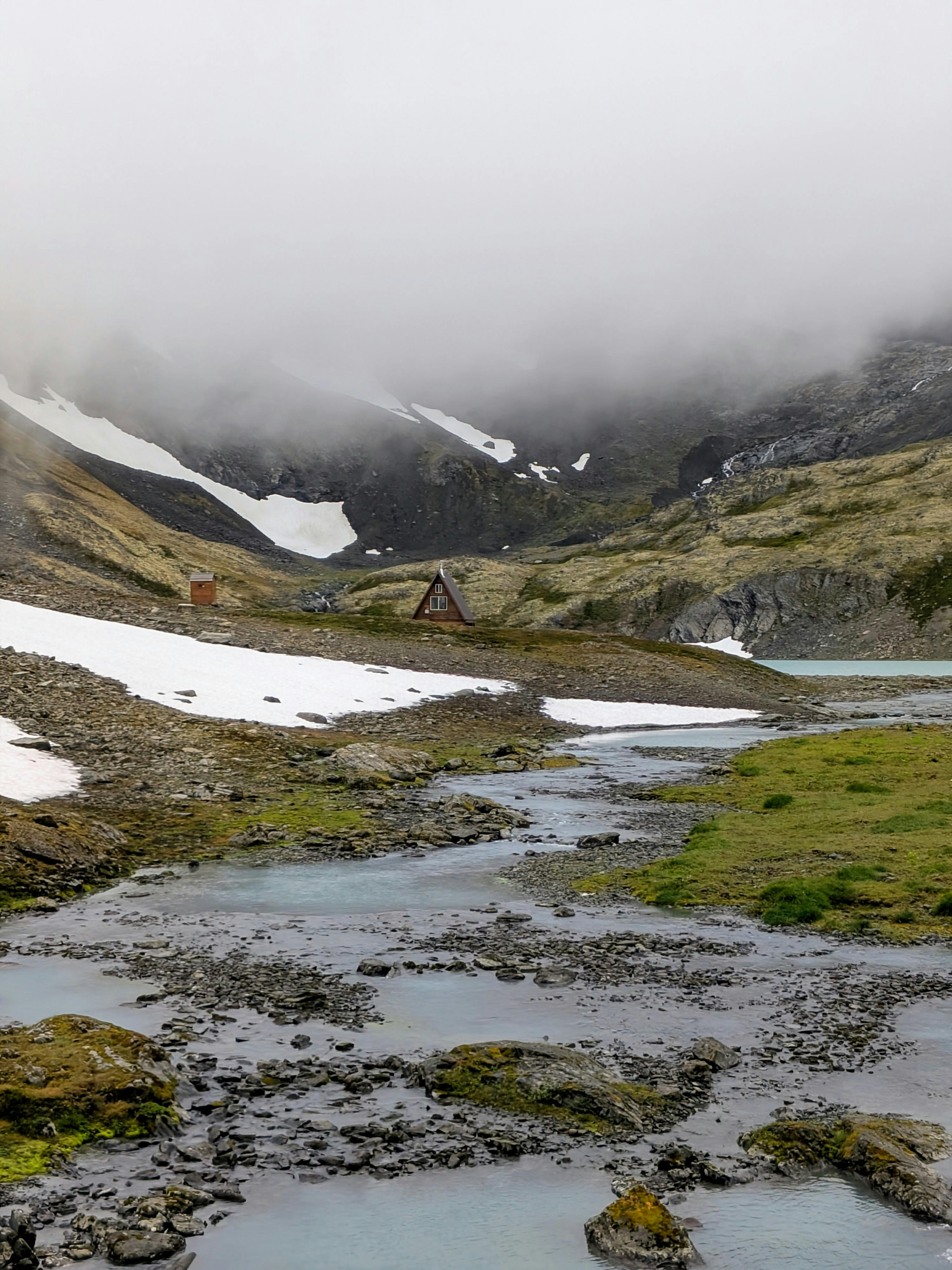 A serene mountainous landscape with a small cabin, stream, and misty peaks.