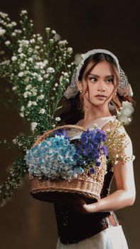 Stylish woman holding basket of white, blue, and purple flowers. Rustic charm, serene mood.
