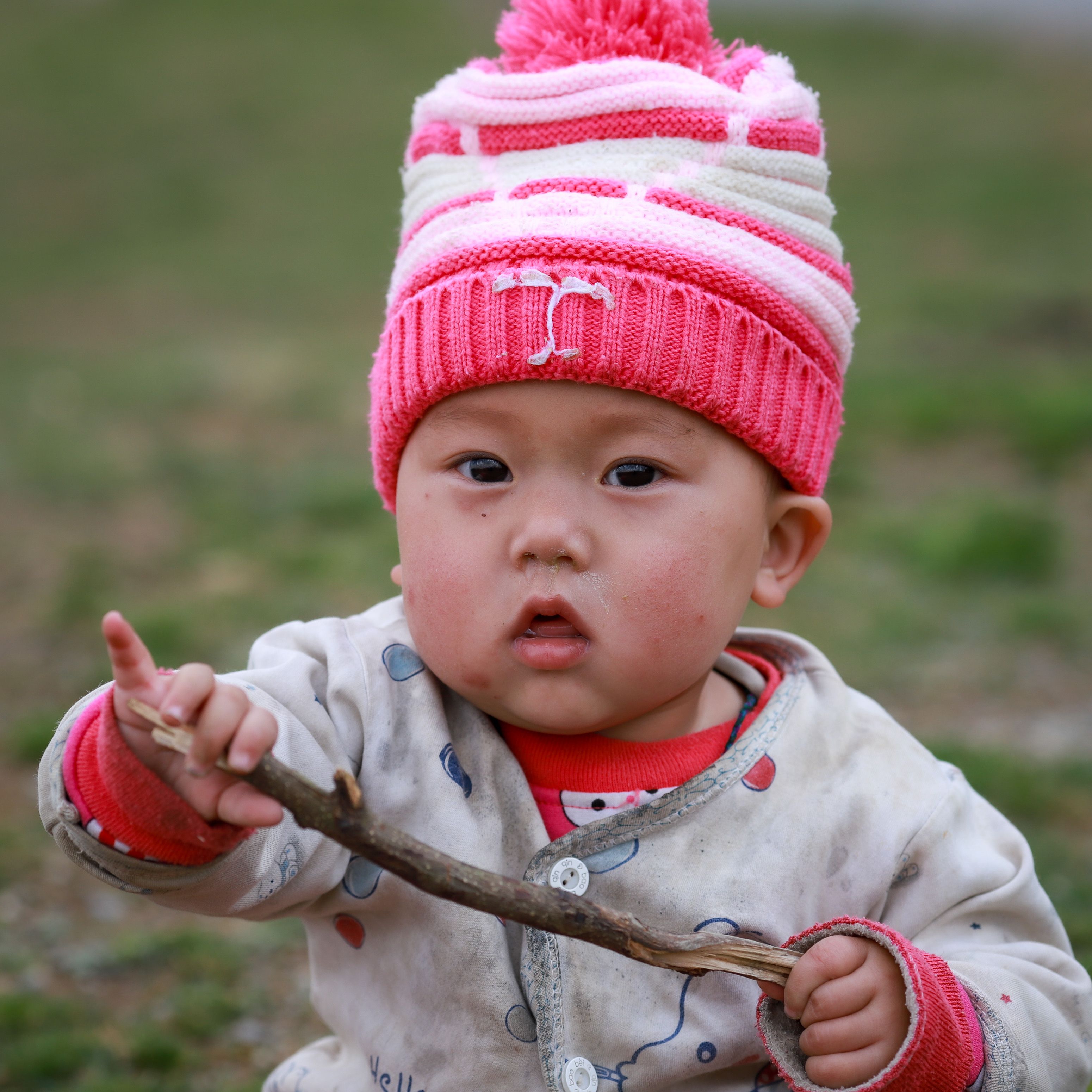 A cute baby wearing a colorful hat and jacket, holding a stick outdoors in Lào Cai, Vietnam.