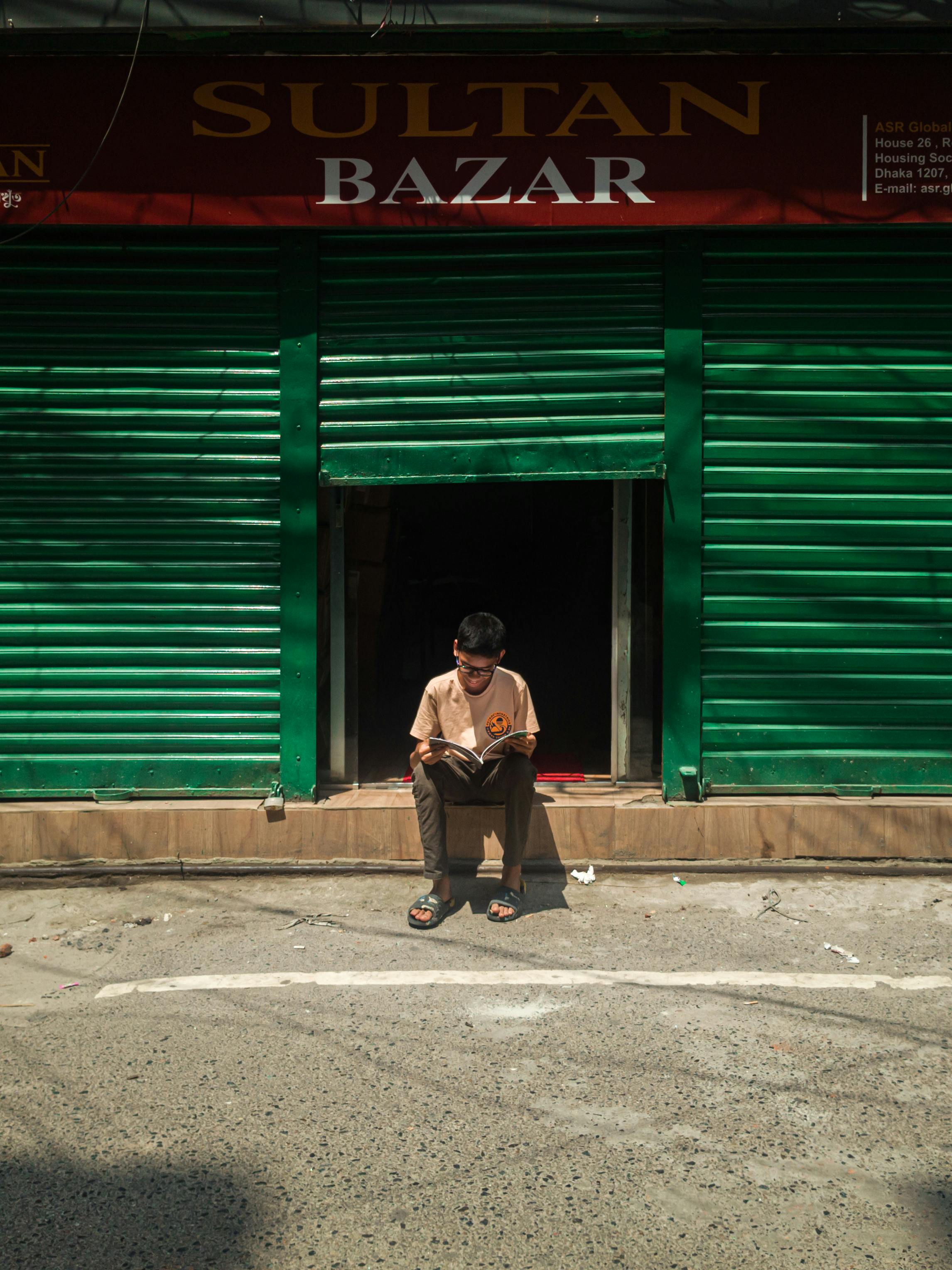 A young boy engrossed in a book outside Sultan Bazar, Dhaka, embodying urban life and reading culture.