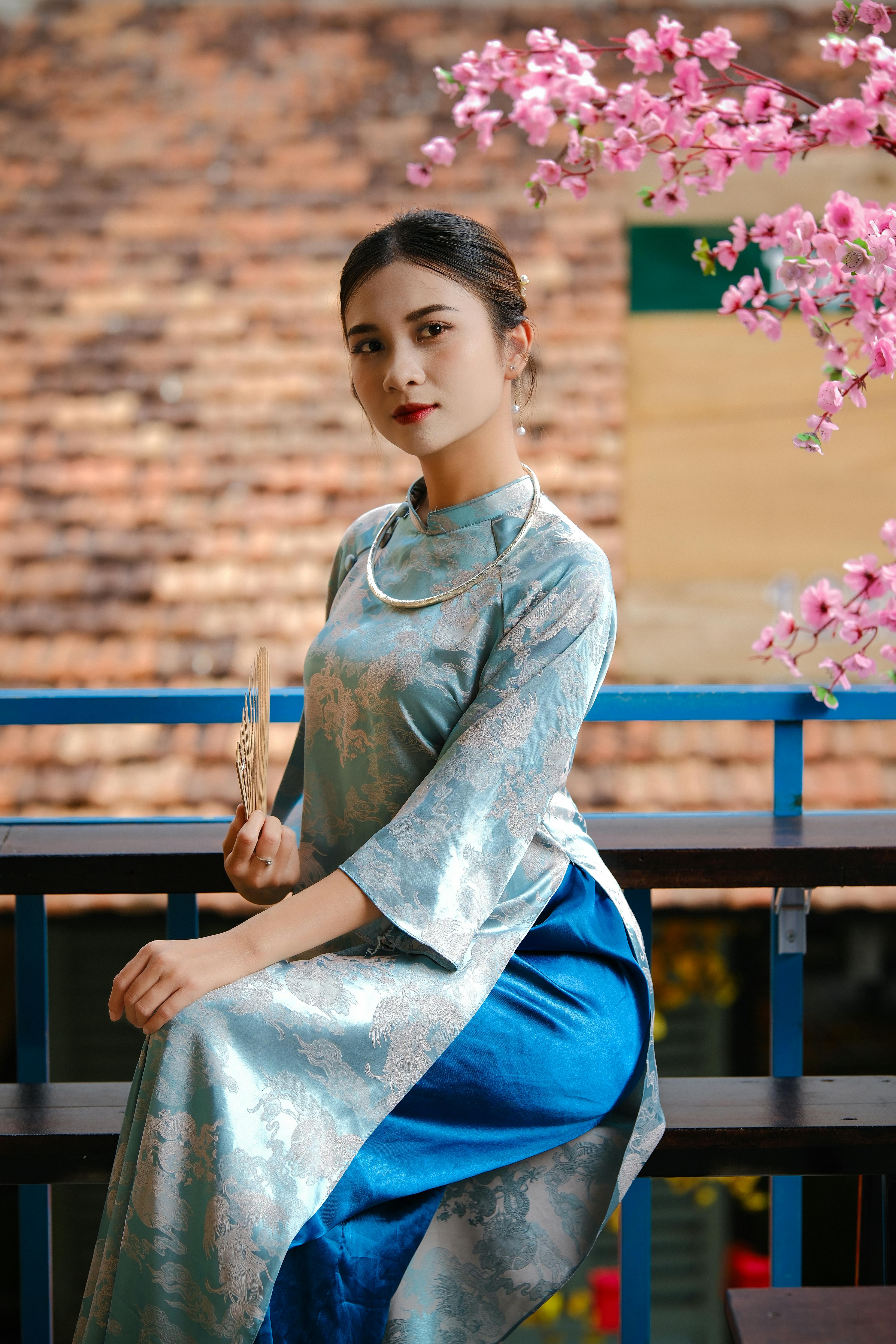 Woman in a traditional elegant dress holding a fan, surrounded by cherry blossoms.