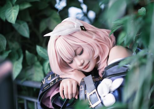 A cosplayer with pink hair rests among lush green foliage outdoors.
