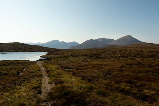 A tranquil Scottish Highlands scene with mountains, loch, and moorland under clear skies.