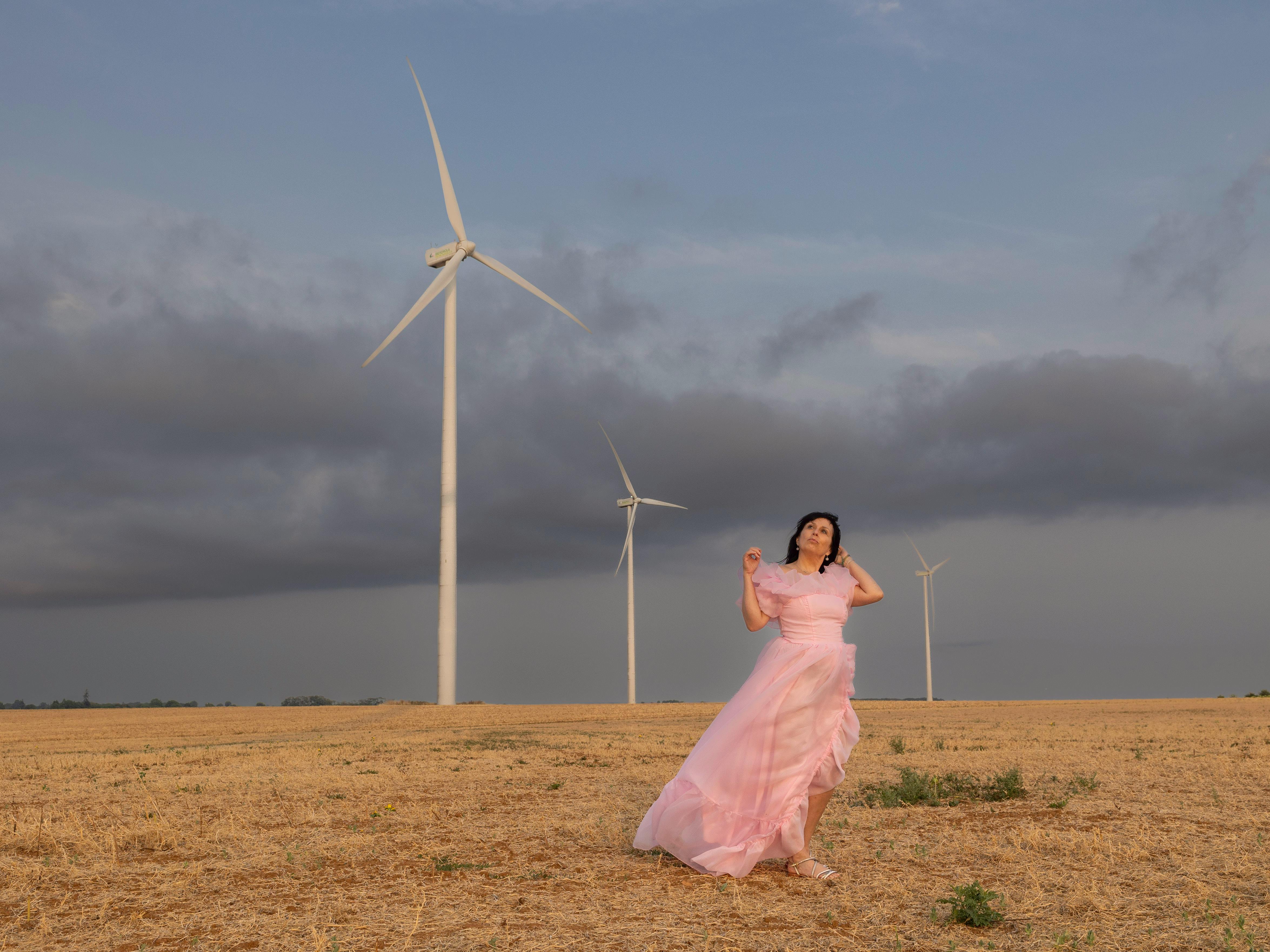 A woman in a flowing pink dress poses gracefully in a field with wind turbines in Pamproux, France. guide