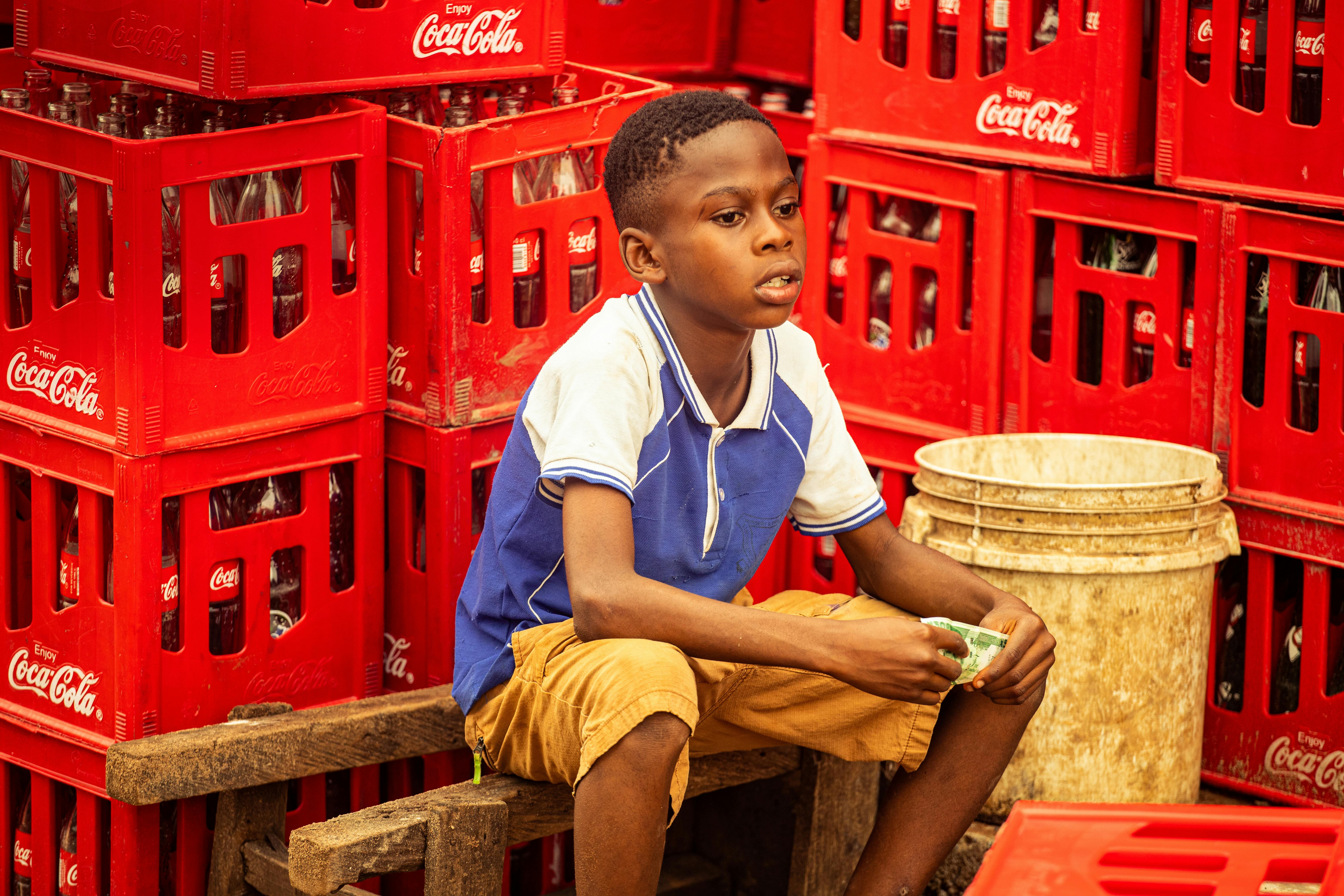 Gratis Vivace foto di strada di un ragazzino seduto tra casse rosse di Coca-Cola ad Abuja, Nigeria. Foto a disposizione
