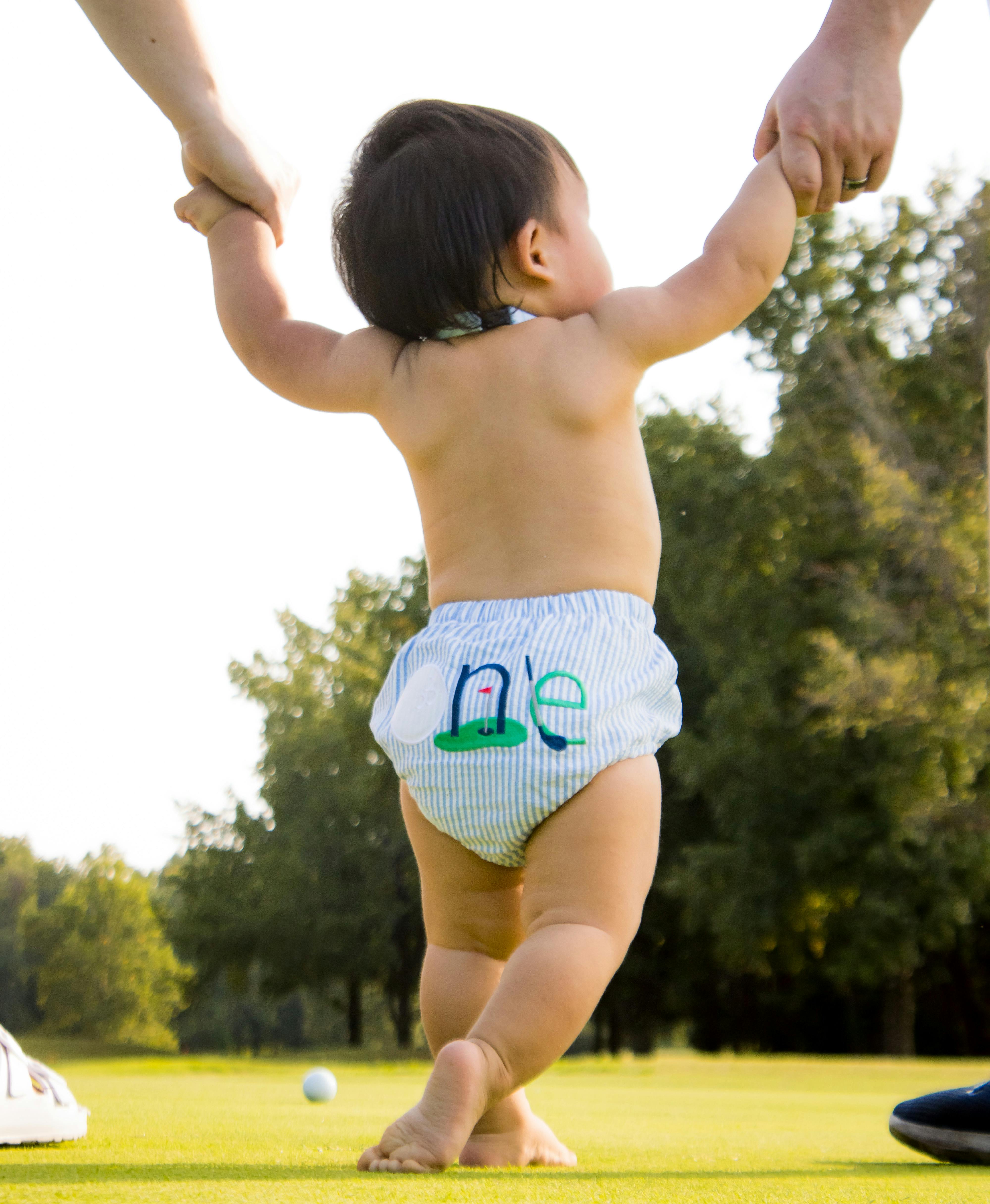 A baby takes first steps on grass, supported by parent hands.