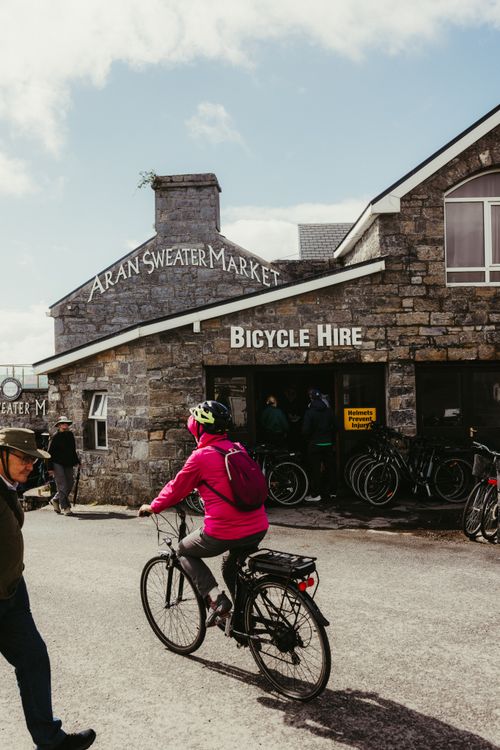 Free Cyclist in pink jacket rides past the Aran Sweater Market on Inishmore, County Galway, Ireland. Stock Photo
