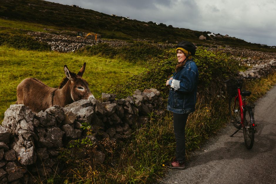 A woman interacts with a donkey near a stone wall on Inishmore, Galway, Ireland, with a bicycle beside her.