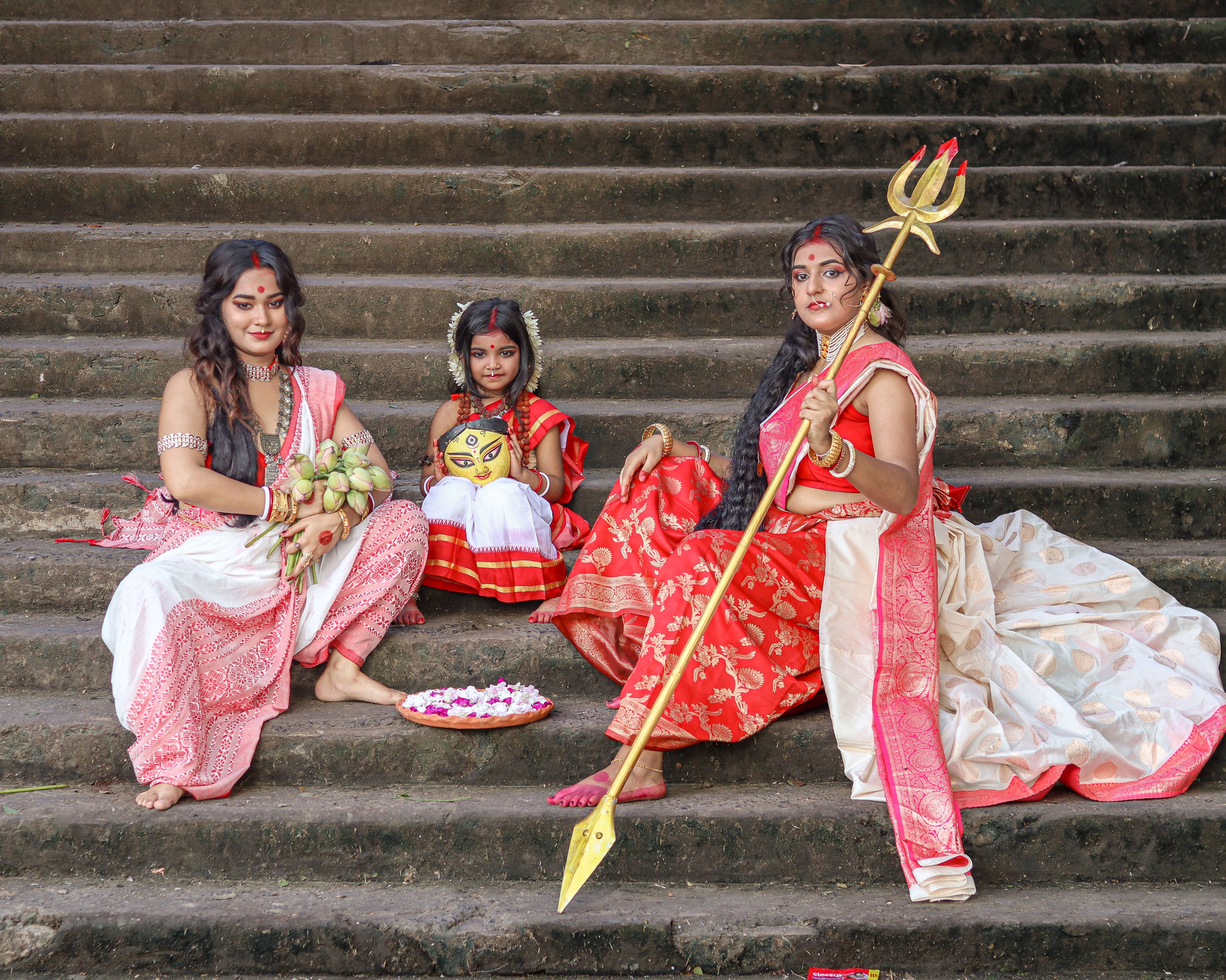 Three women in traditional Indian attire sit on stone steps with cultural items.