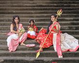 Three women in traditional Indian attire sit on stone steps with cultural items.