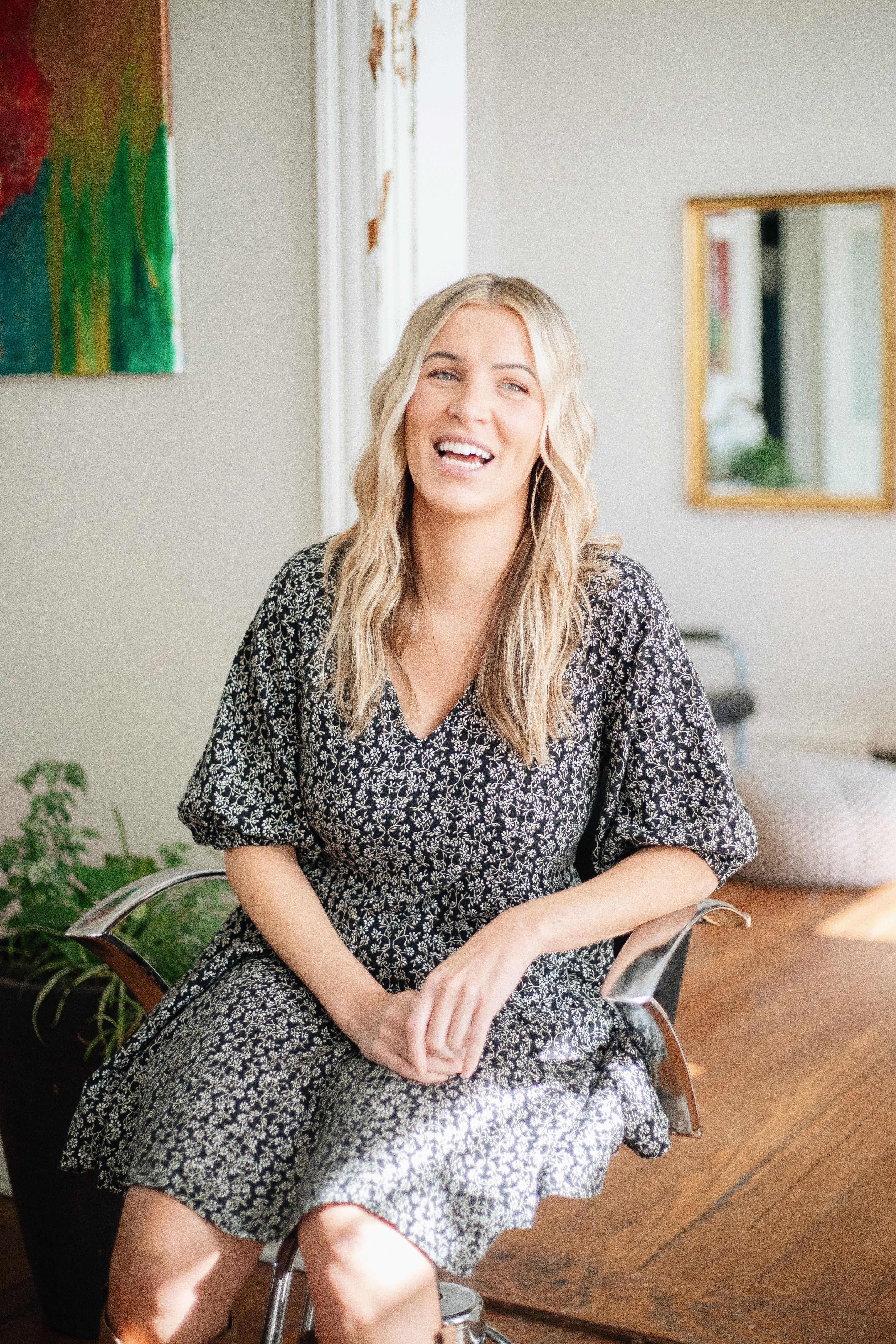 Natural light portrait of a smiling woman with wavy blonde hair in a stylish salon setting.