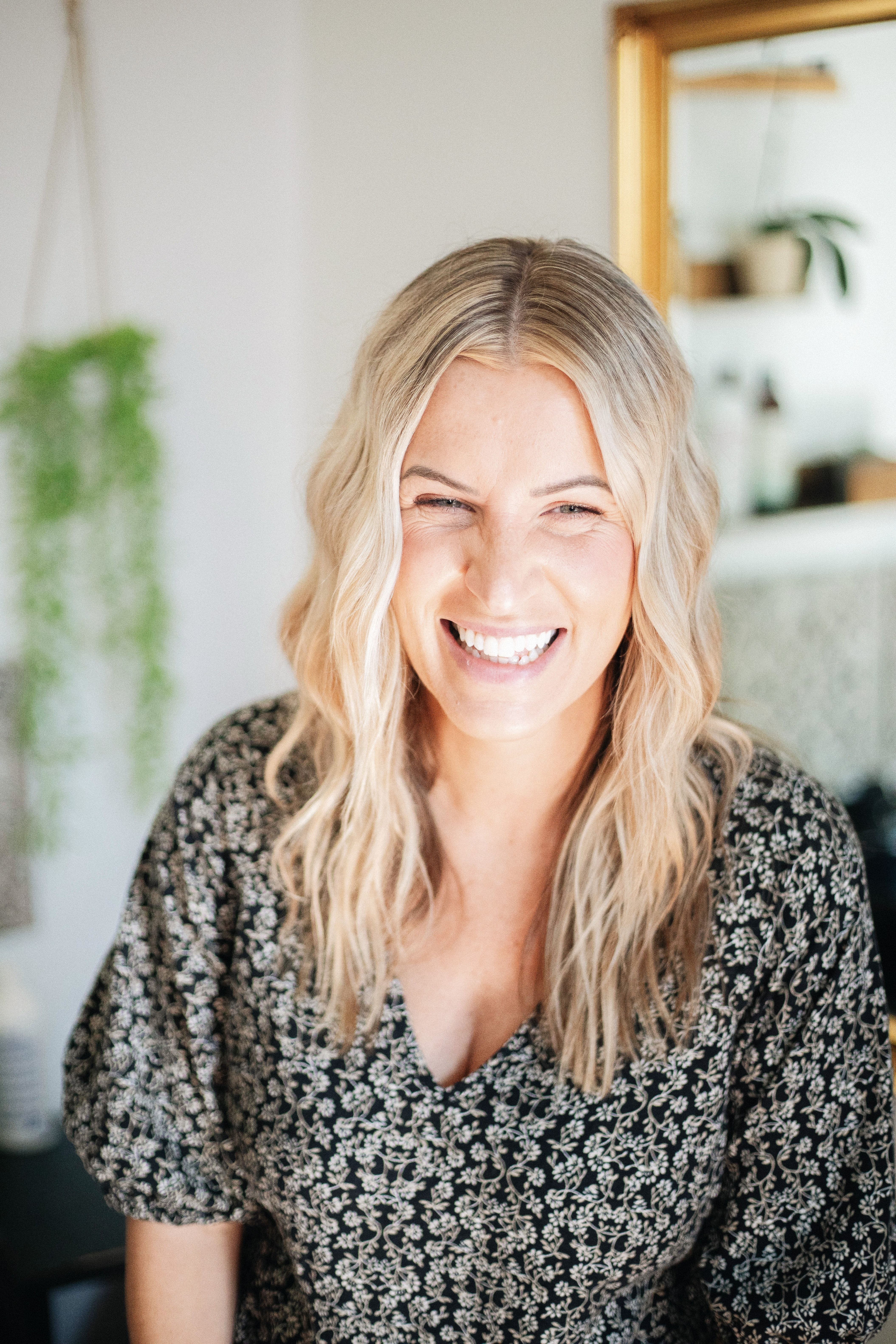 A joyful woman with blonde wavy hair smiling in natural light indoors.