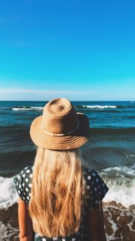 A woman in a straw hat and polka dot dress stands by the ocean, enjoying a sunny beach view.