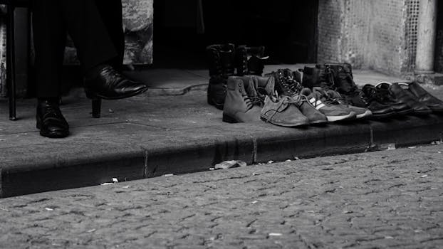 Monochrome photo of various shoes lined up on a curb with a person's foot visible, evoking street photography charm.