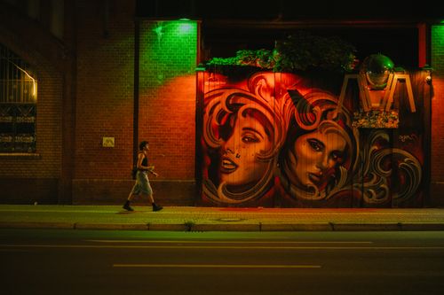 Free A person walks past colorful street art murals at night under vibrant street lights. Stock Photo
