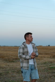 A man in casual attire stands in a field at dusk, looking pensive against a serene sky.