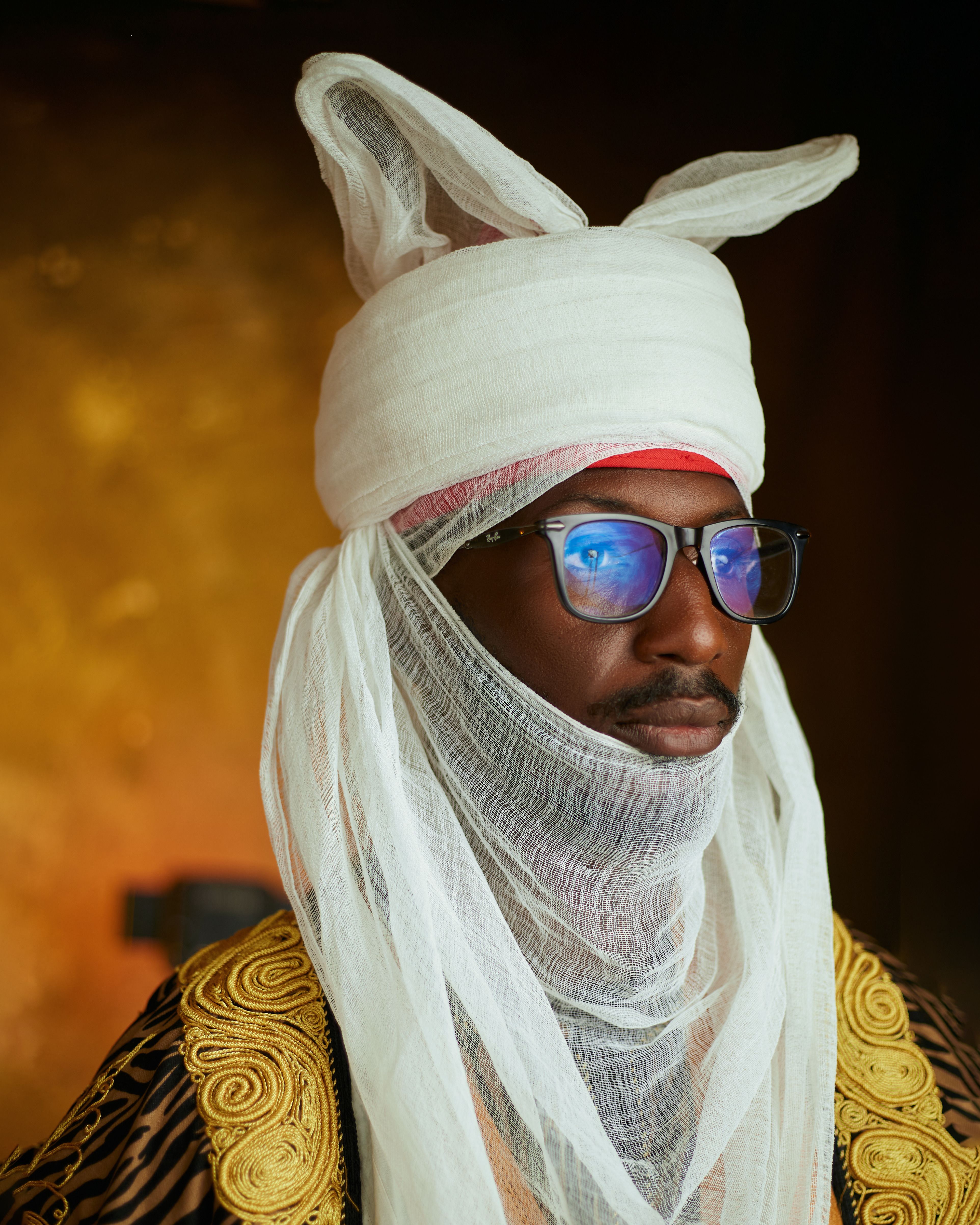 Man in traditional African attire with glasses, veiled face in warm setting.