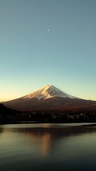 Serene image of Mount Fuji at sunset with a crescent moon above, mirrored on a tranquil lake.