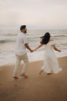 A couple in white clothes walking hand in hand along a sandy beach at sunset.
